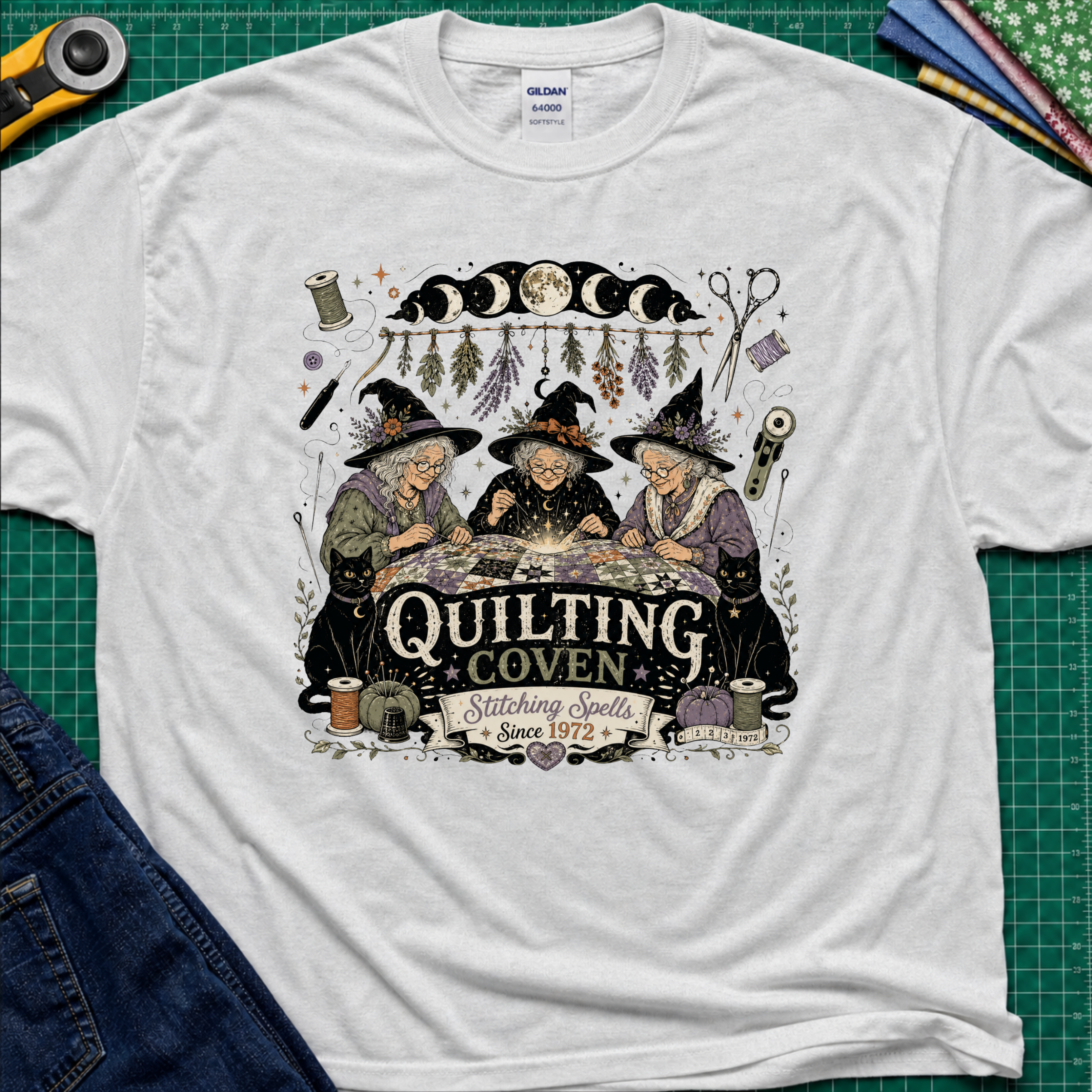 A light grey Gildan t-shirt with a detailed graphic of three witches sewing at a table under a moon phase design, with the text 'Quilting Coven Stitching Spells Since 1972'. The shirt is laid flat on a green cutting mat with craft tools and fabric visible around it.