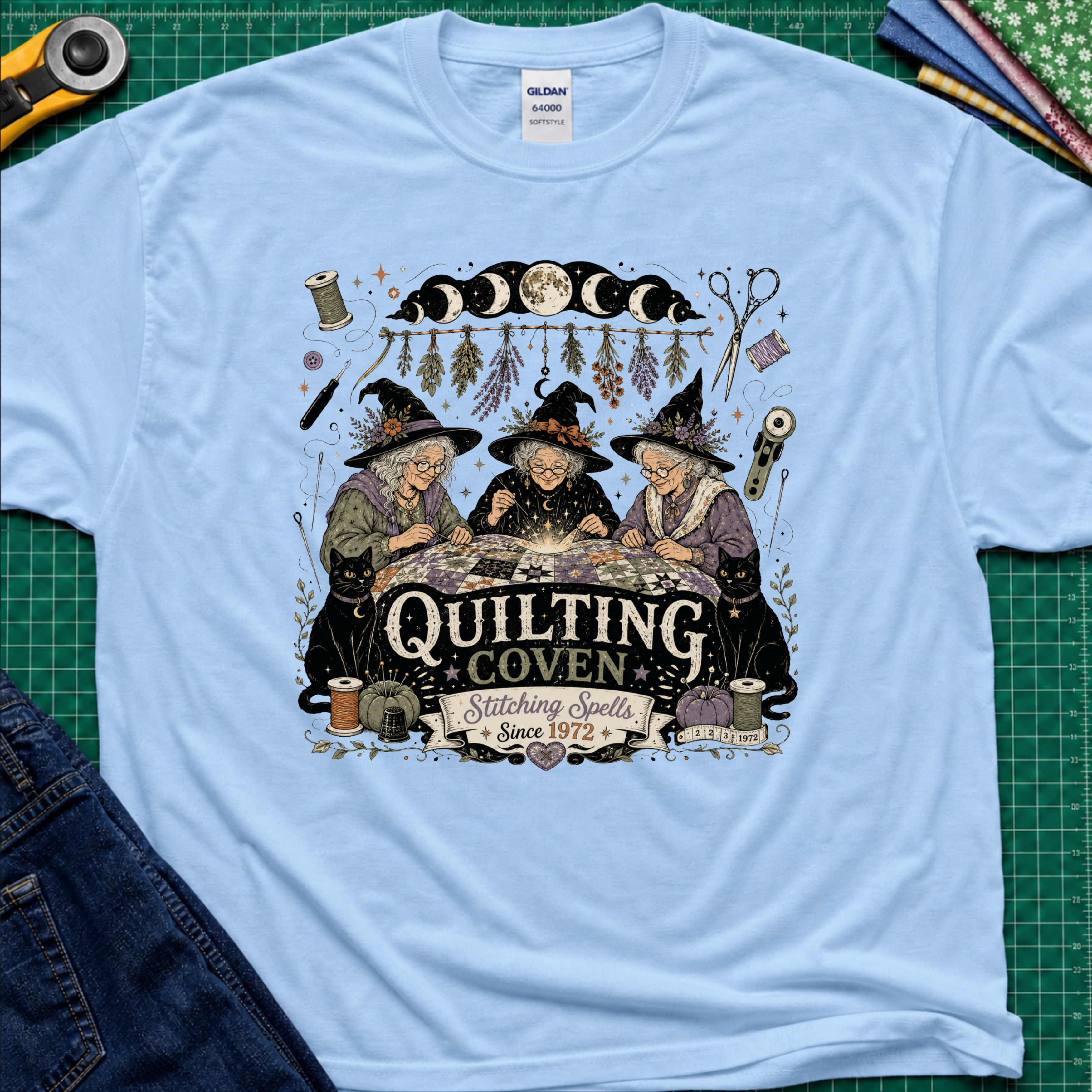 A light blue Gildan t-shirt with a detailed graphic of three witches sewing at a table under a moon phase design, with the text 'Quilting Coven Stitching Spells Since 1972'. The shirt is laid flat on a green cutting mat with craft tools and fabric visible around it.