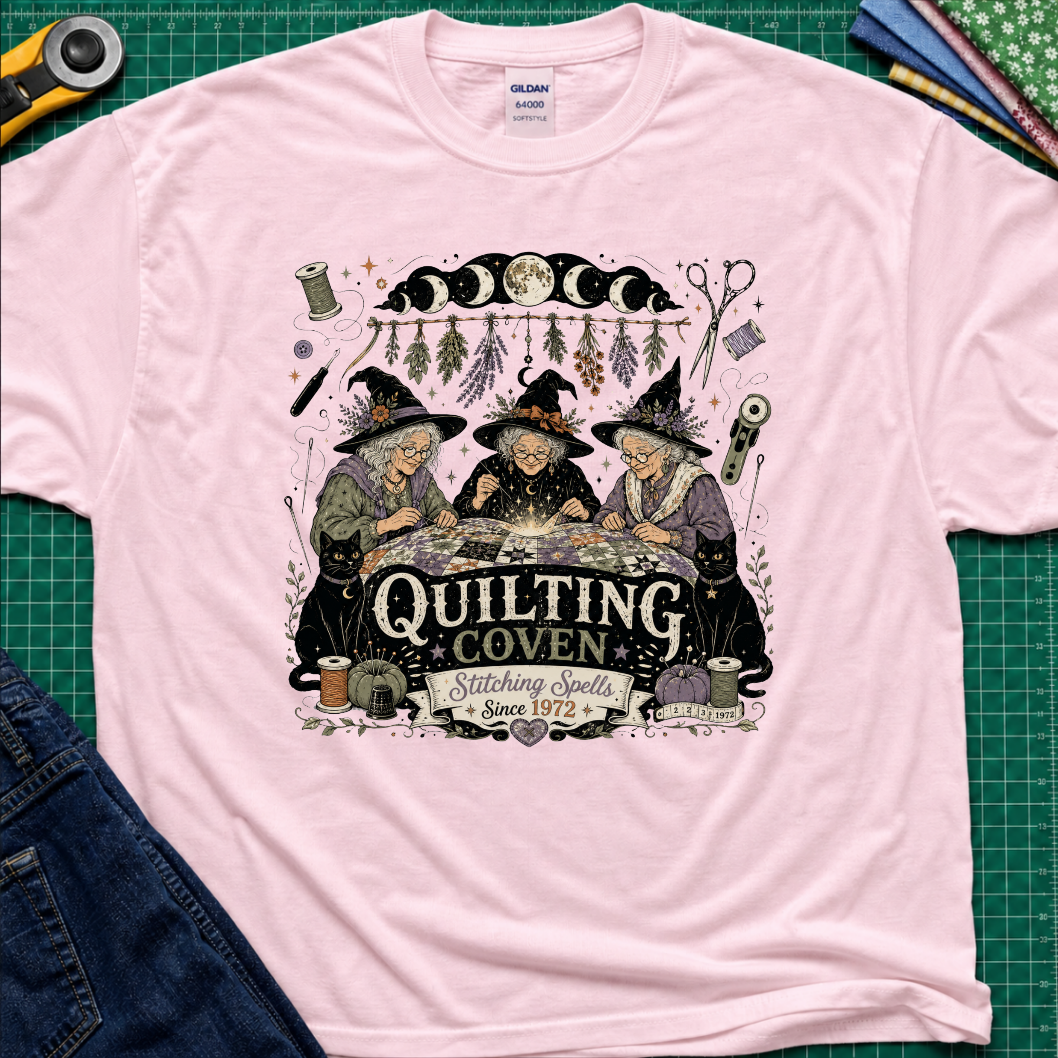 A light pink Gildan t-shirt with a detailed graphic of three witches sewing at a table under a moon phase design, with the text 'Quilting Coven Stitching Spells Since 1972'. The shirt is laid flat on a green cutting mat with craft tools and fabric visible around it.
