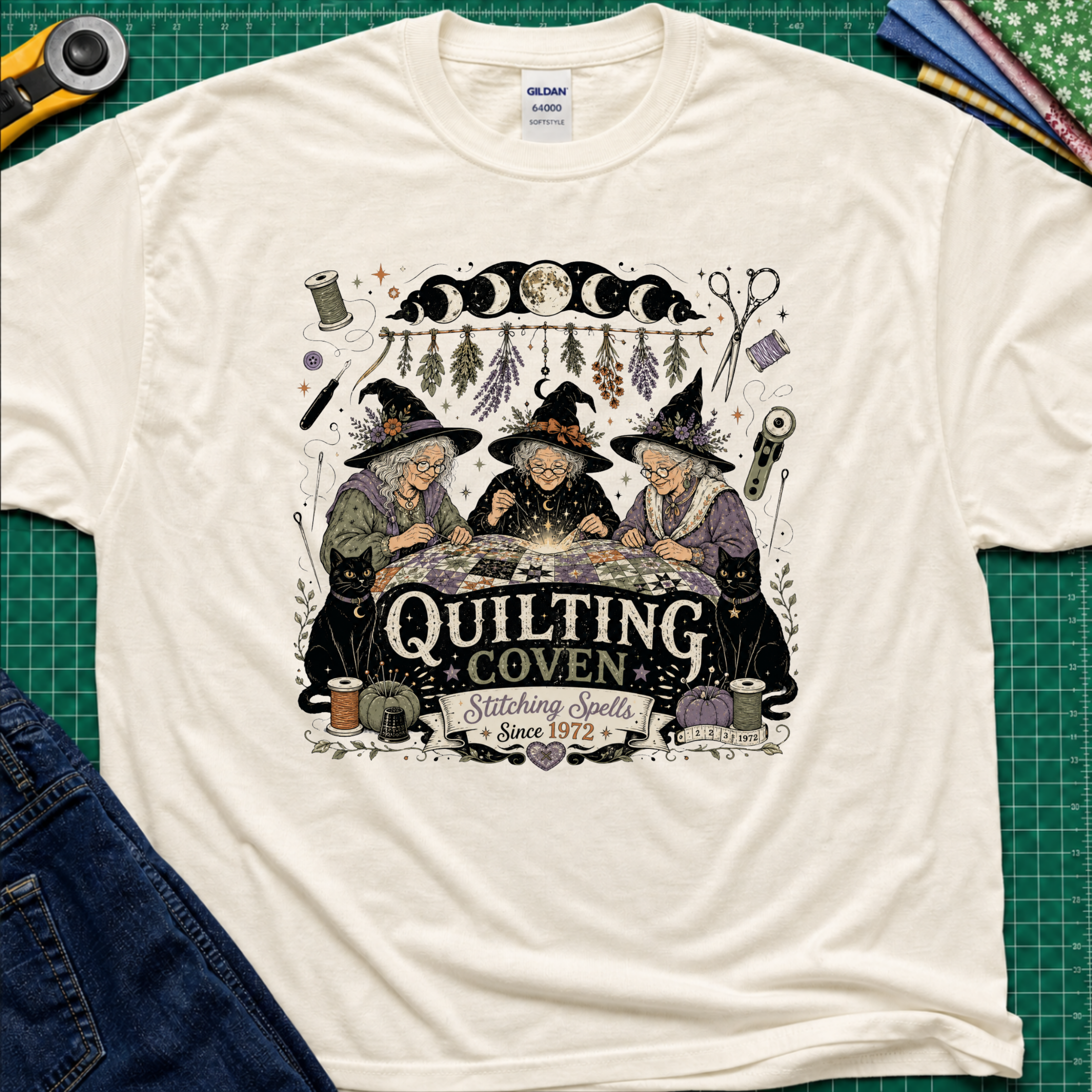 A light cream Gildan t-shirt with a detailed graphic of three witches sewing at a table under a moon phase design, with the text 'Quilting Coven Stitching Spells Since 1972'. The shirt is laid flat on a green cutting mat with craft tools and fabric visible around it.