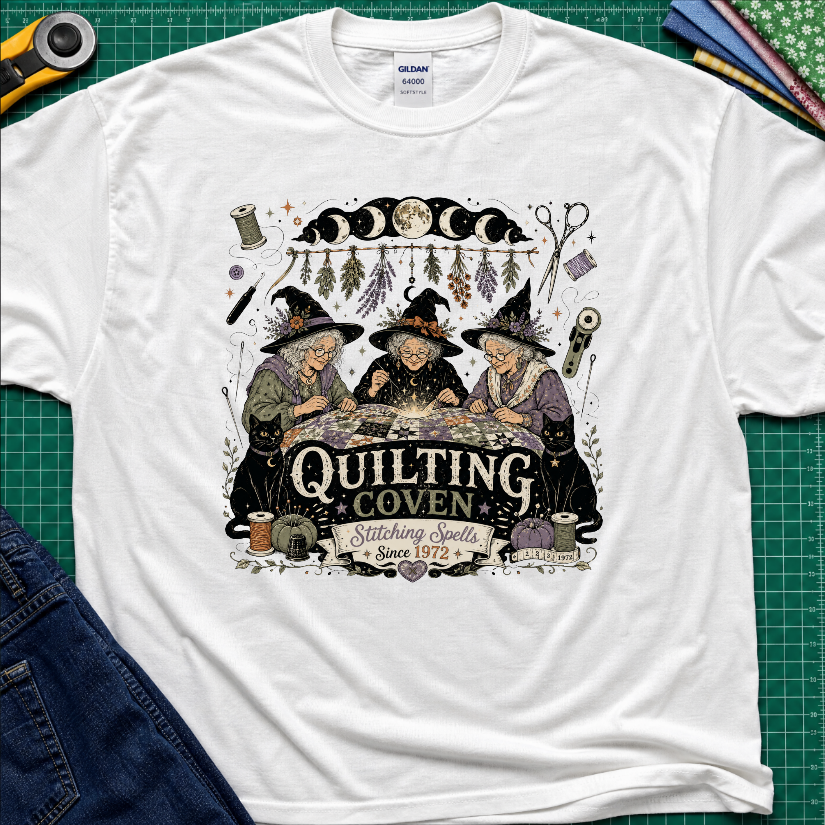 A white Gildan t-shirt with a detailed graphic of three witches sewing at a table under a moon phase design, with the text 'Quilting Coven Stitching Spells Since 1972'. The shirt is laid flat on a green cutting mat with craft tools and fabric visible around it.