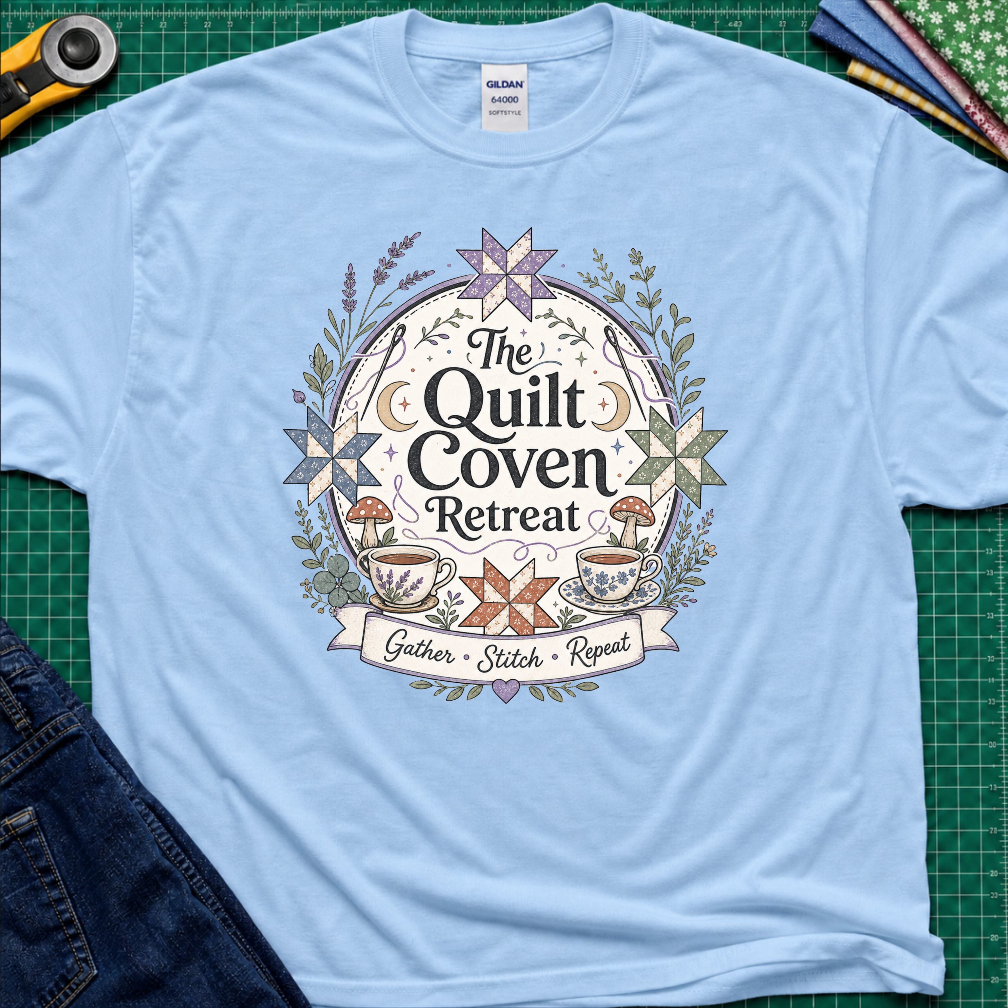 A light blue Gildan t-shirt with a circular graphic design reading 'The Quilt Coven Retreat' surrounded by patchwork stars, leaves, and coffee cups, laid flat on a green cutting mat with sewing supplies.