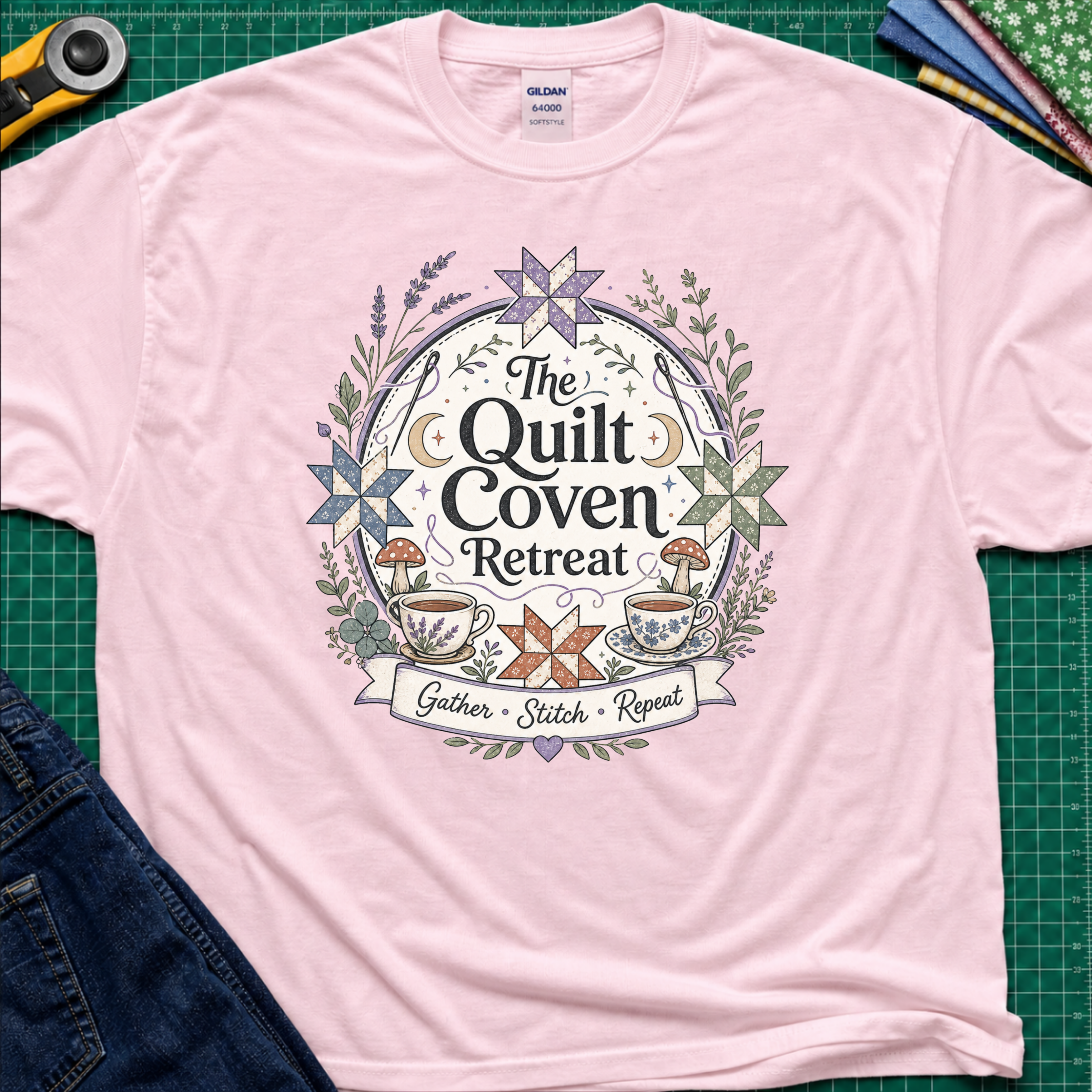 A light pink Gildan t-shirt with a circular graphic design reading 'The Quilt Coven Retreat' surrounded by patchwork stars, leaves, and coffee cups, laid flat on a green cutting mat with sewing supplies.