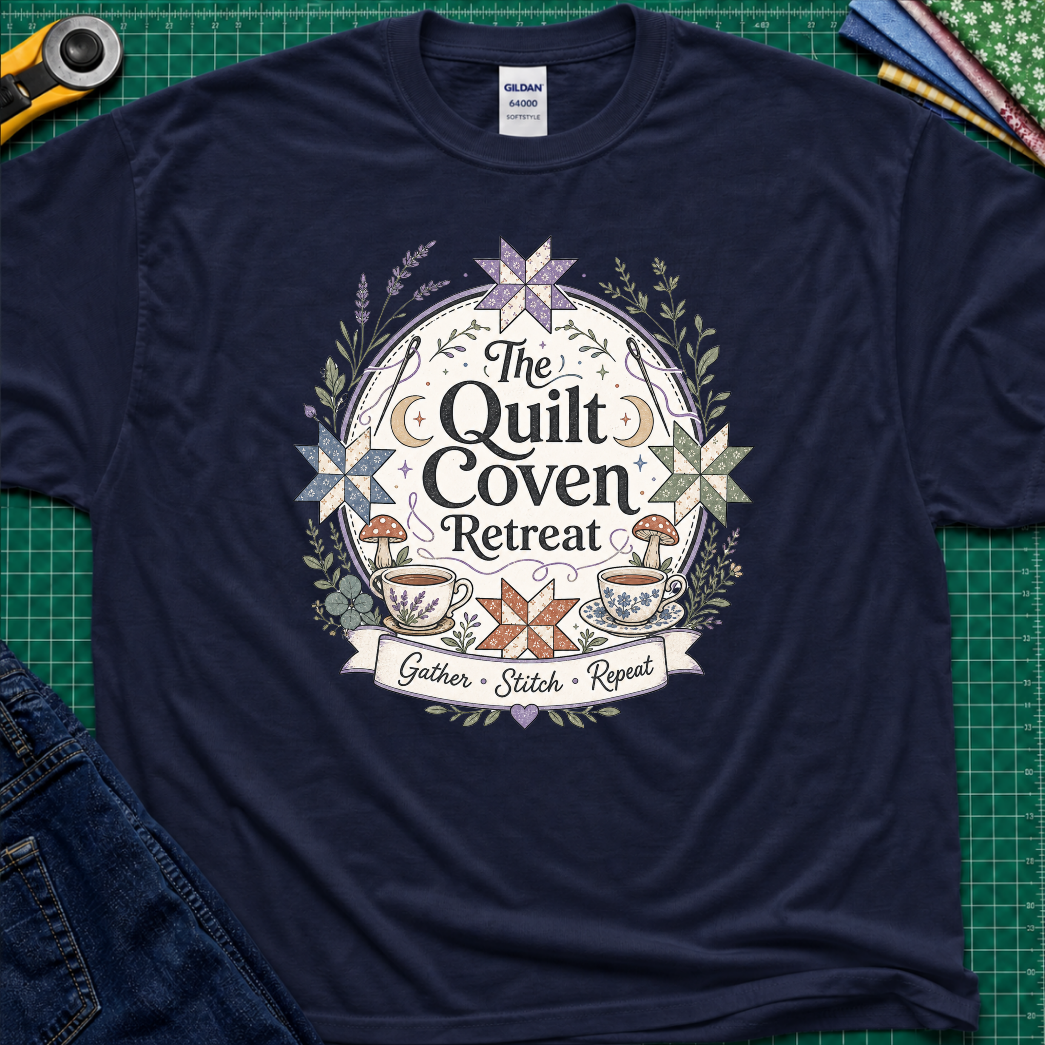 A navy blue Gildan t-shirt with a circular graphic design reading 'The Quilt Coven Retreat' surrounded by patchwork stars, leaves, and coffee cups, laid flat on a green cutting mat with sewing supplies.
