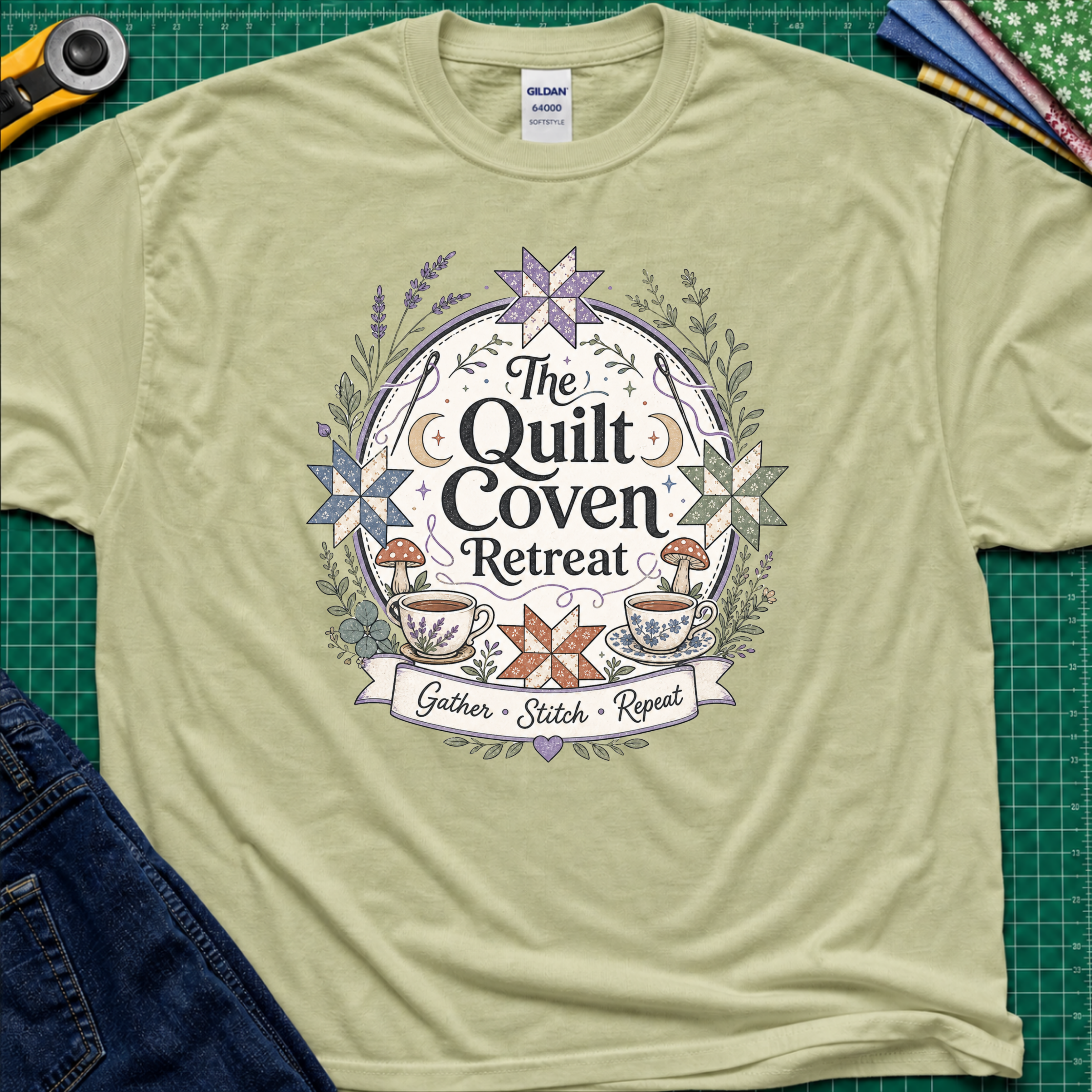 A light pistachio green Gildan t-shirt with a circular graphic design reading 'The Quilt Coven Retreat' surrounded by patchwork stars, leaves, and coffee cups, laid flat on a green cutting mat with sewing supplies.