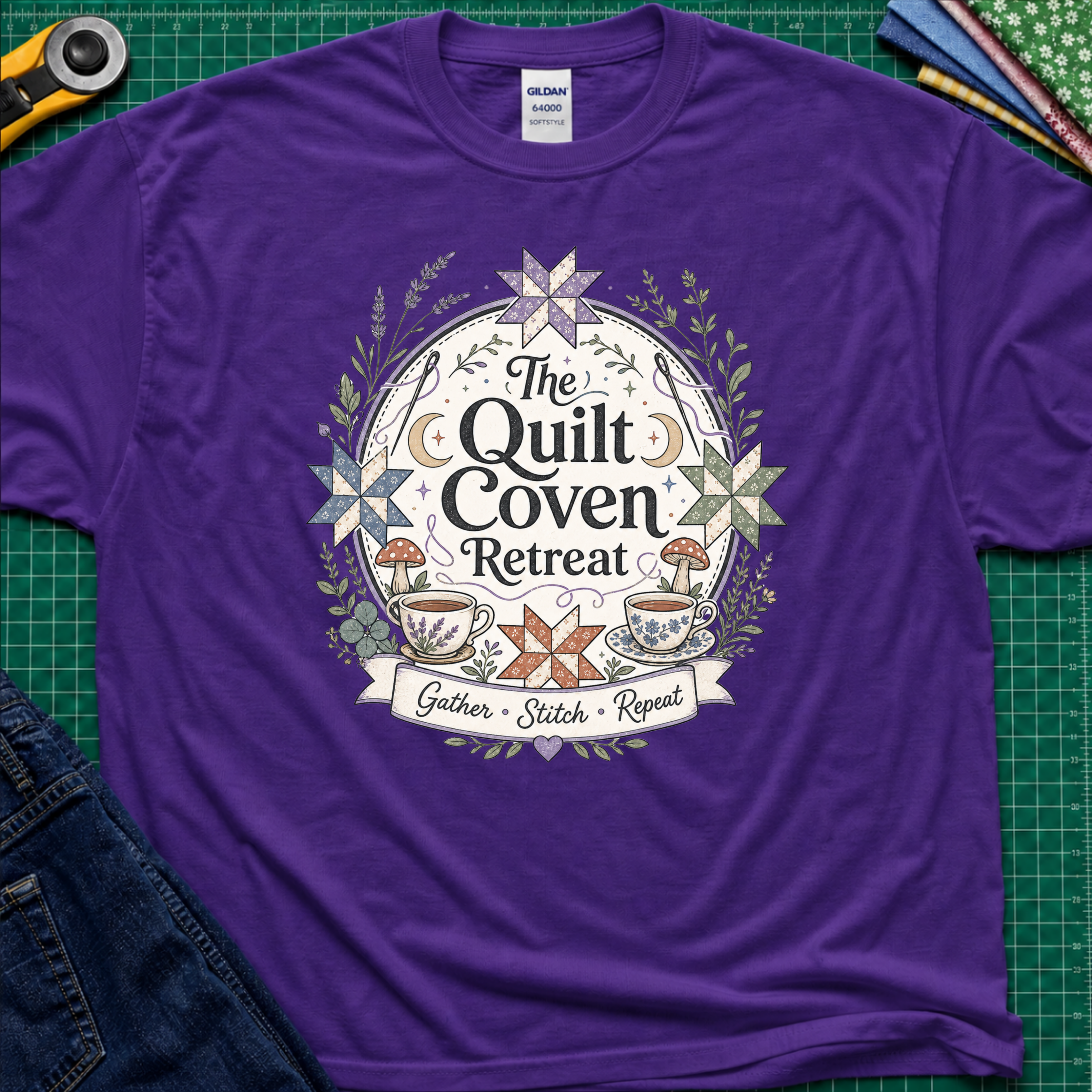 A purple Gildan t-shirt with a circular graphic design reading 'The Quilt Coven Retreat' surrounded by patchwork stars, leaves, and coffee cups, laid flat on a green cutting mat with sewing supplies.