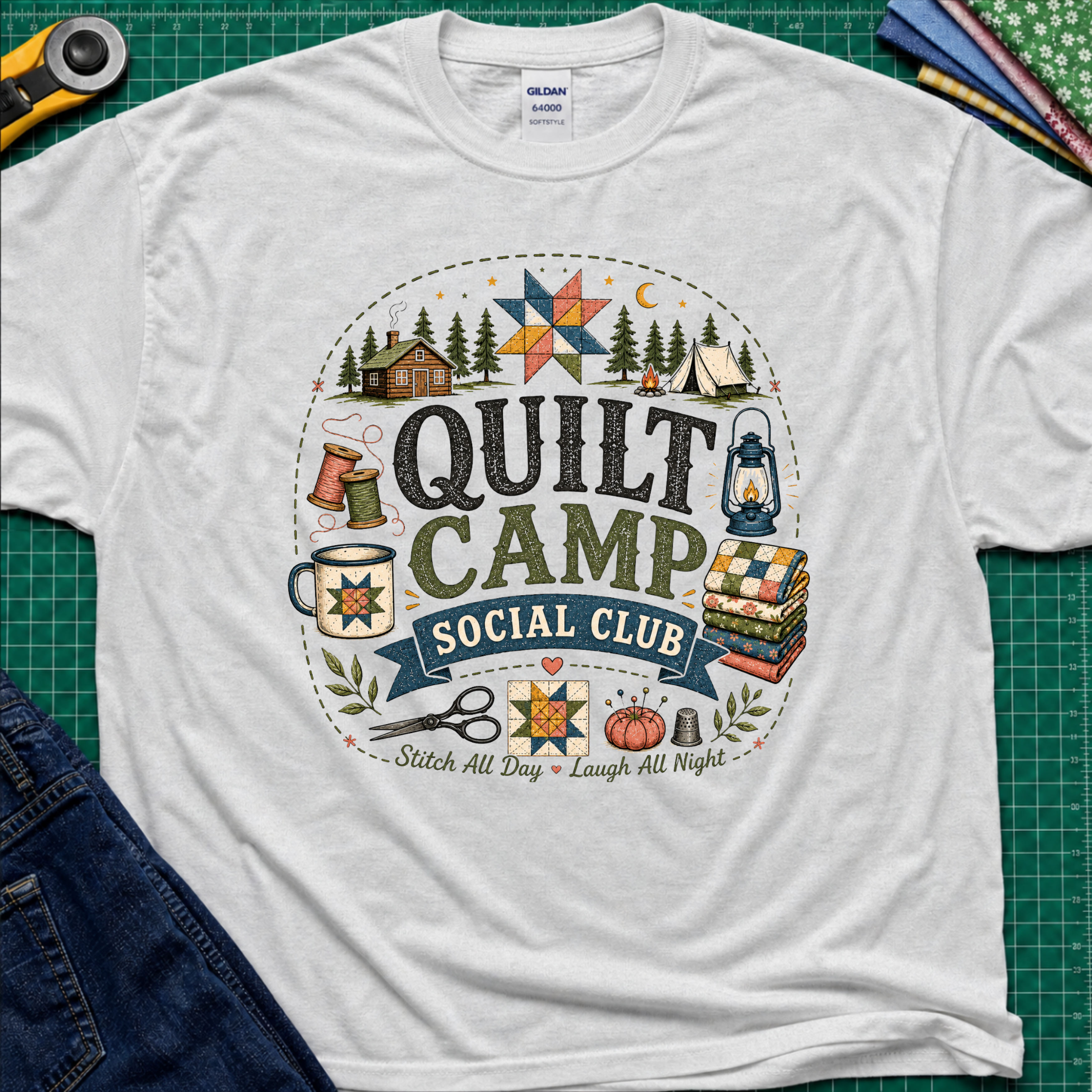 A light grey Gildan t-shirt featuring a circular 'Quilt Camp Social Club' graphic with quilting and camping illustrations. T-Shirt lays flat on a green cutting mat with a rotary cutter, fabric stack, and denim jeans partically visible in the corners.