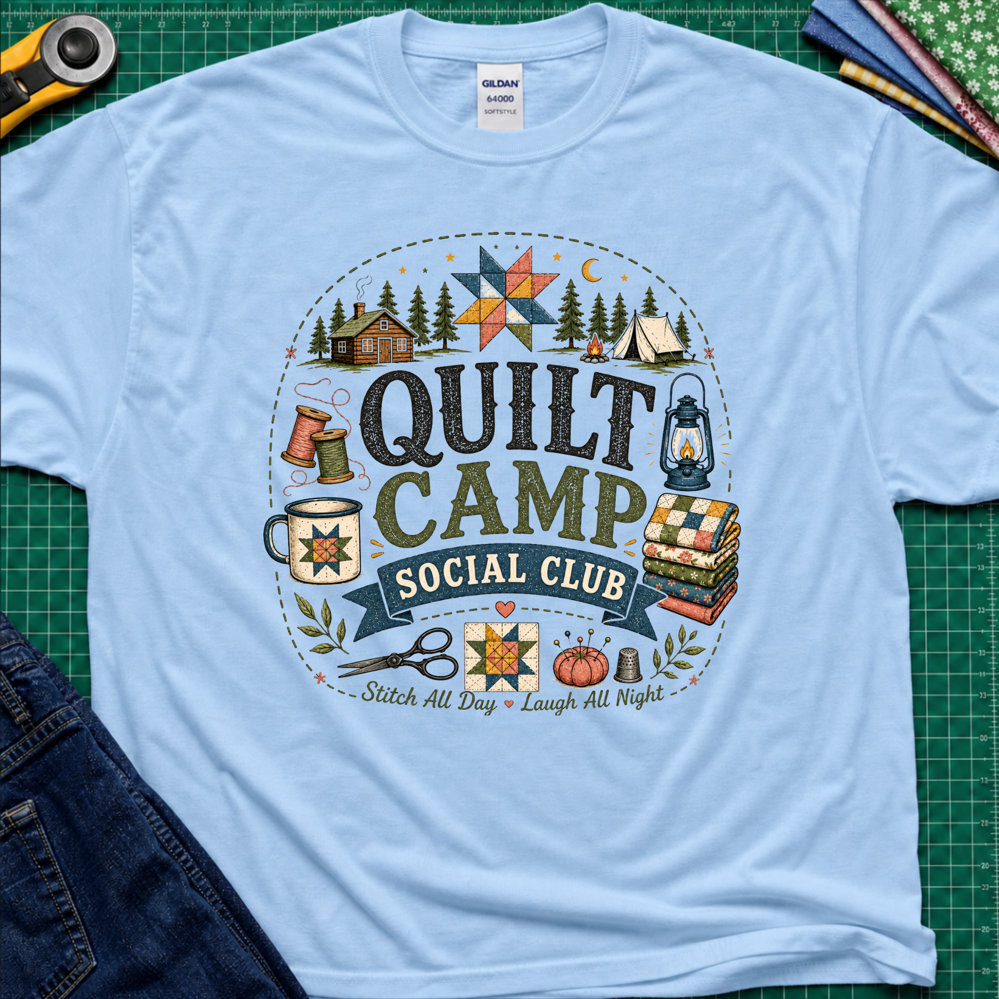 A light blue Gildan t-shirt featuring a circular 'Quilt Camp Social Club' graphic with quilting and camping illustrations. T-Shirt lays flat on a green cutting mat with a rotary cutter, fabric stack, and denim jeans partically visible in the corners.