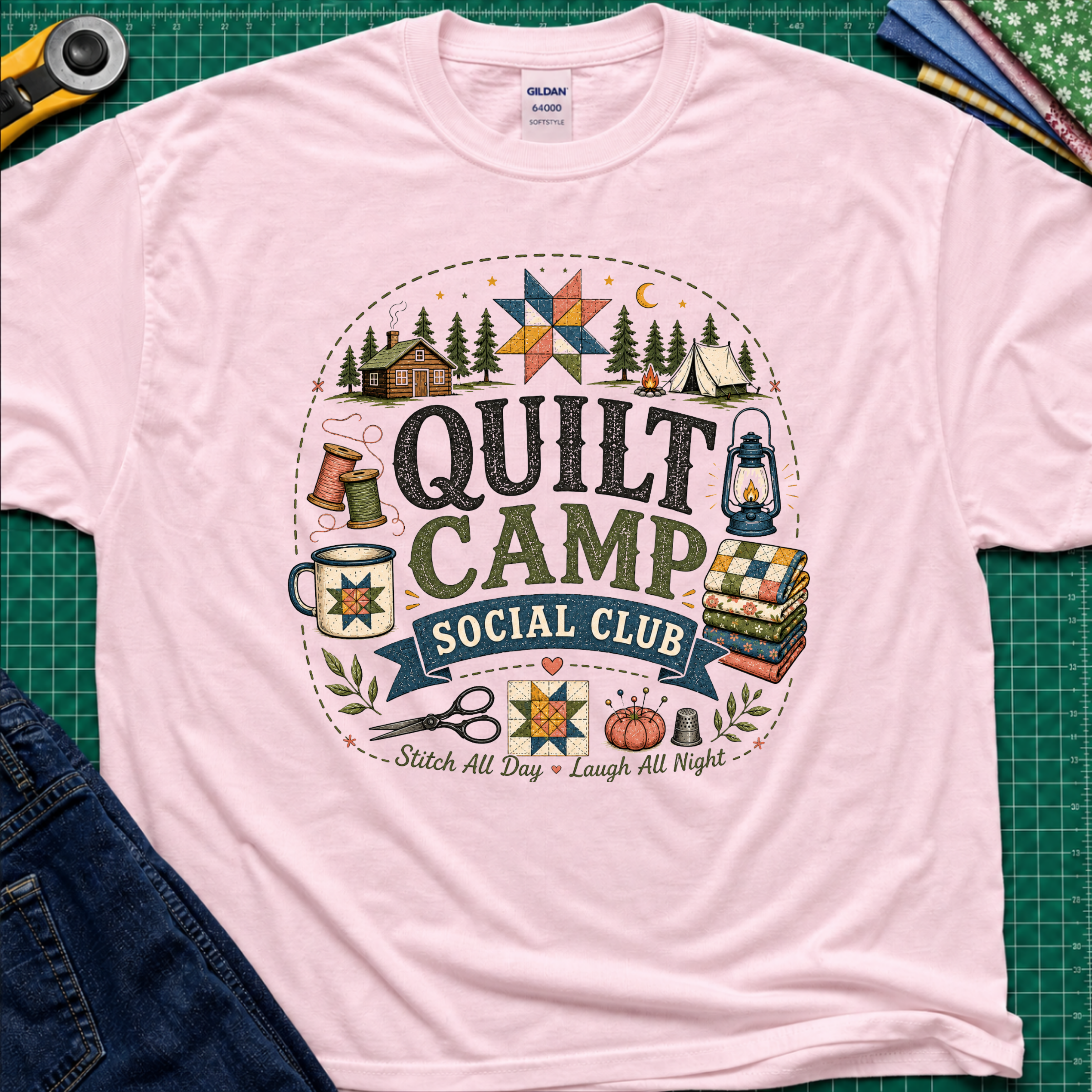 A light pink Gildan t-shirt featuring a circular 'Quilt Camp Social Club' graphic with quilting and camping illustrations. T-Shirt lays flat on a green cutting mat with a rotary cutter, fabric stack, and denim jeans partically visible in the corners.