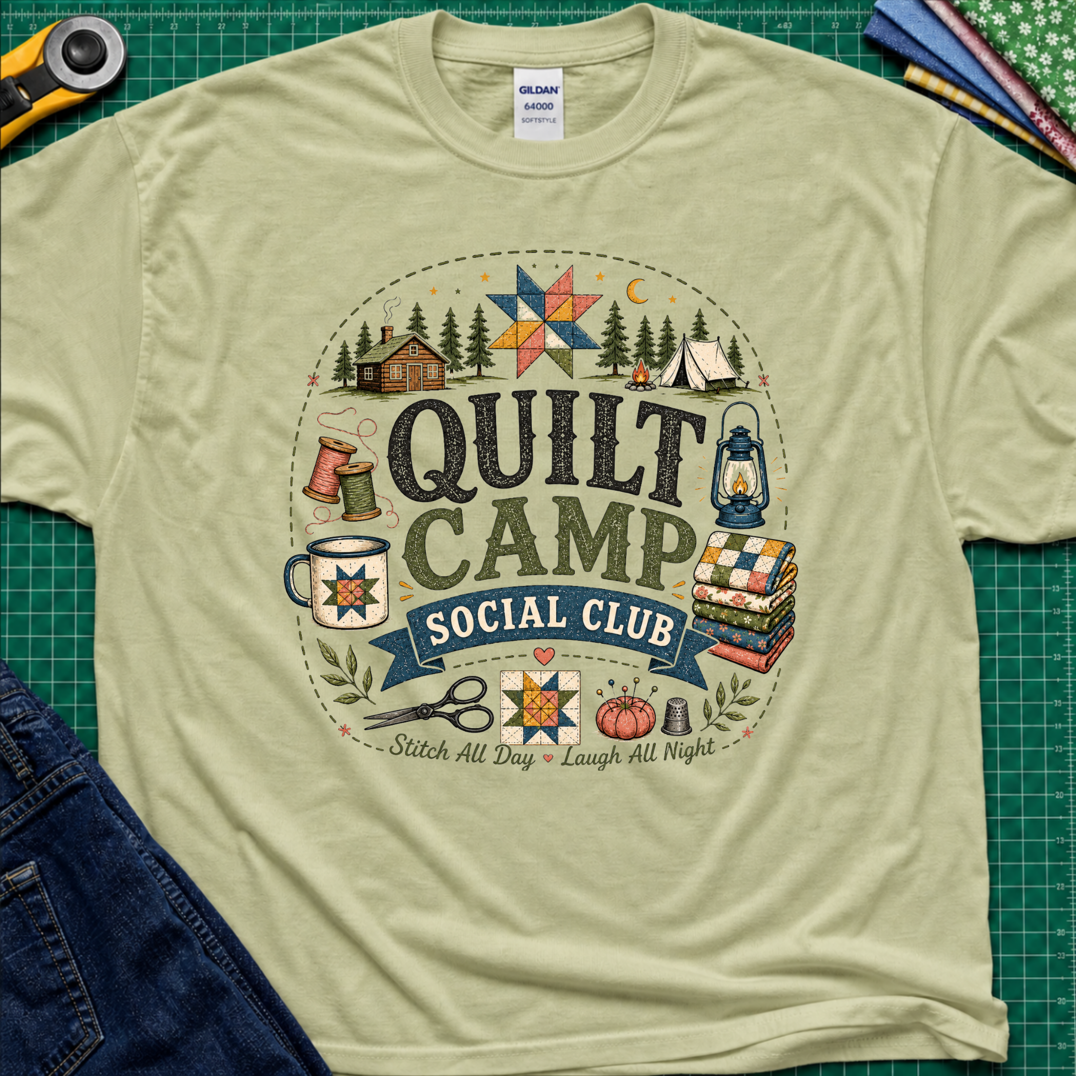 A light pistachio green Gildan t-shirt featuring a circular 'Quilt Camp Social Club' graphic with quilting and camping illustrations. T-Shirt lays flat on a green cutting mat with a rotary cutter, fabric stack, and denim jeans partically visible in the corners.