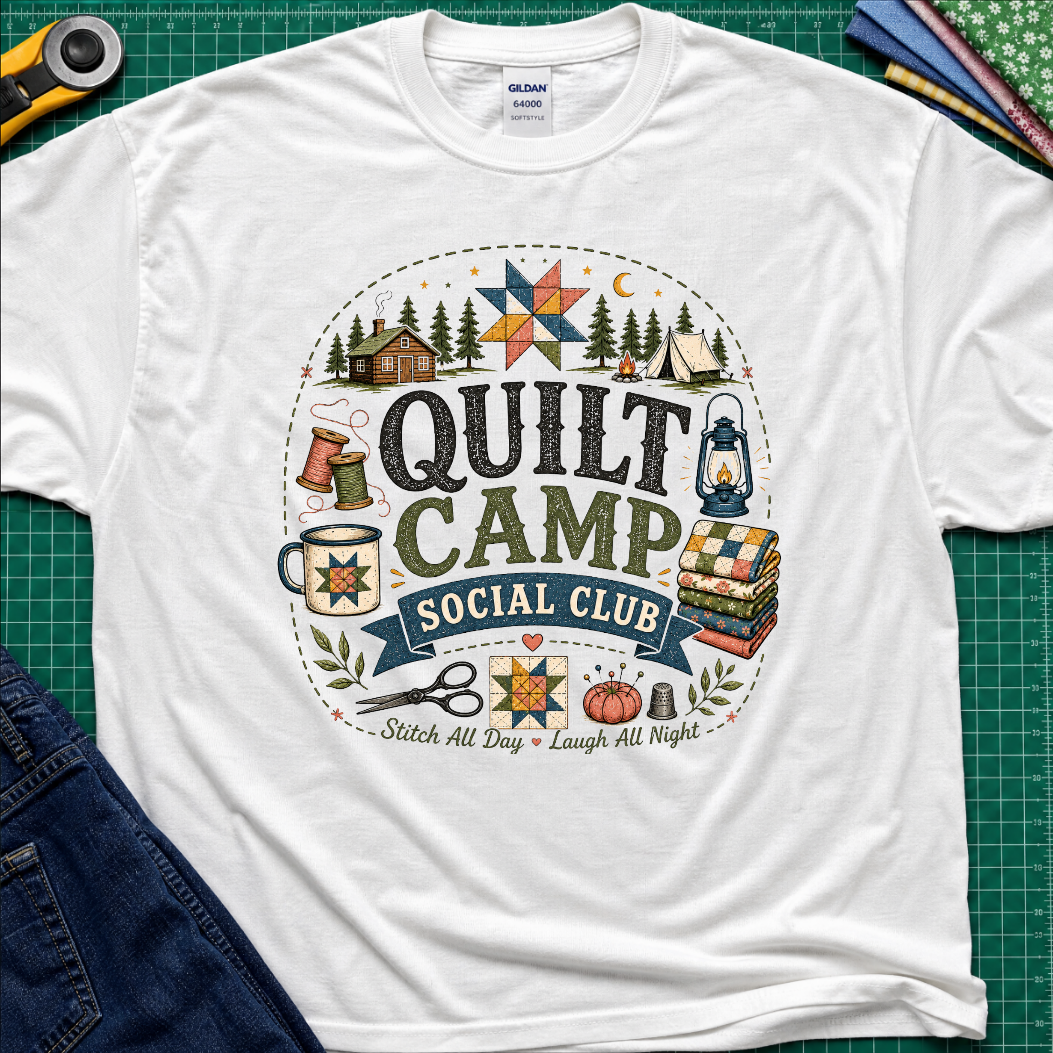 A white Gildan t-shirt featuring a circular 'Quilt Camp Social Club' graphic with quilting and camping illustrations. T-Shirt lays flat on a green cutting mat with a rotary cutter, fabric stack, and denim jeans partically visible in the corners.