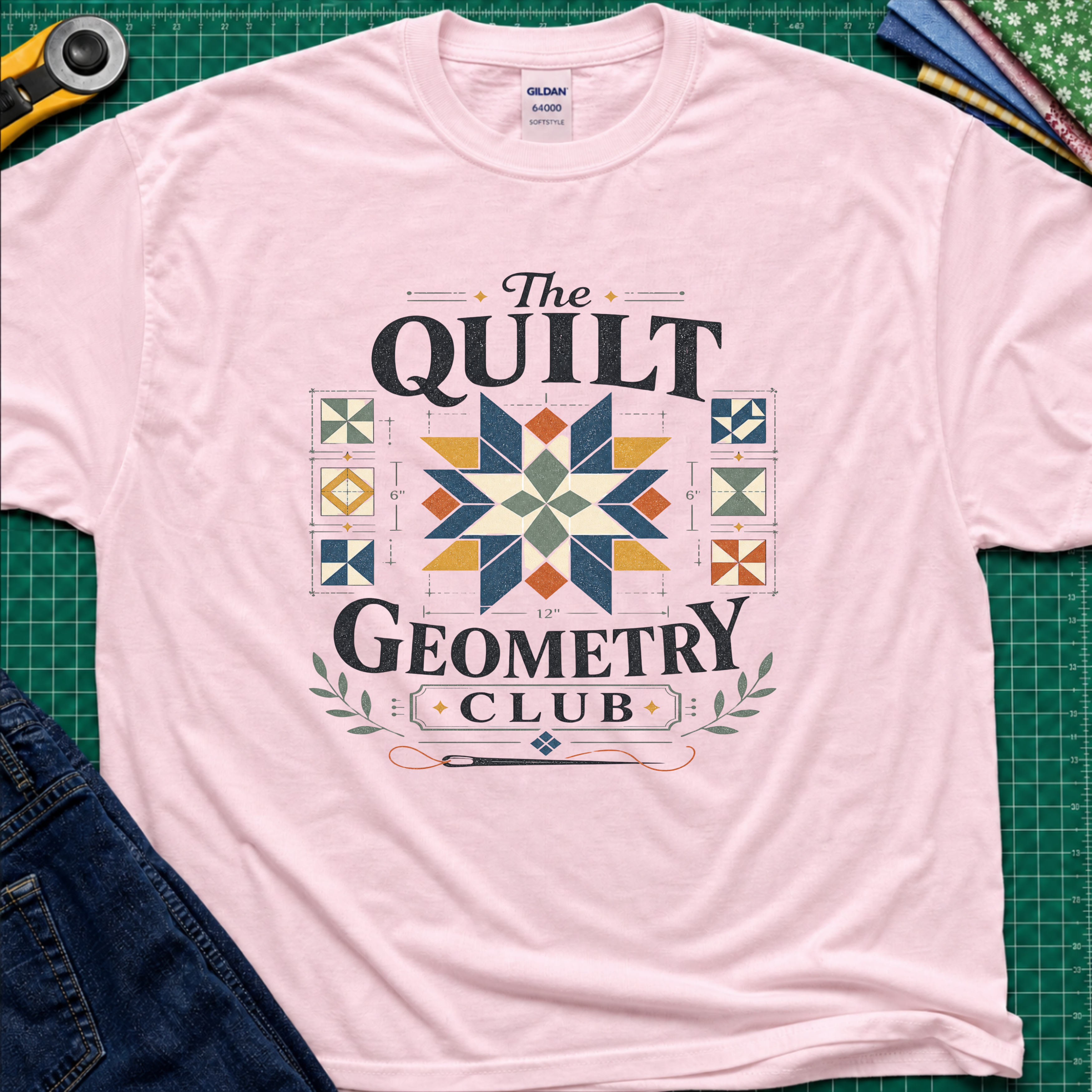 A light pink Gildan t-shirt with the words 'The Quilt Geometry Club' surround a colourful quilt block with smaller quilt blocks on either side. The shirt lays flat on a green cutting mat with craft tools and jeans visible around the edges.
