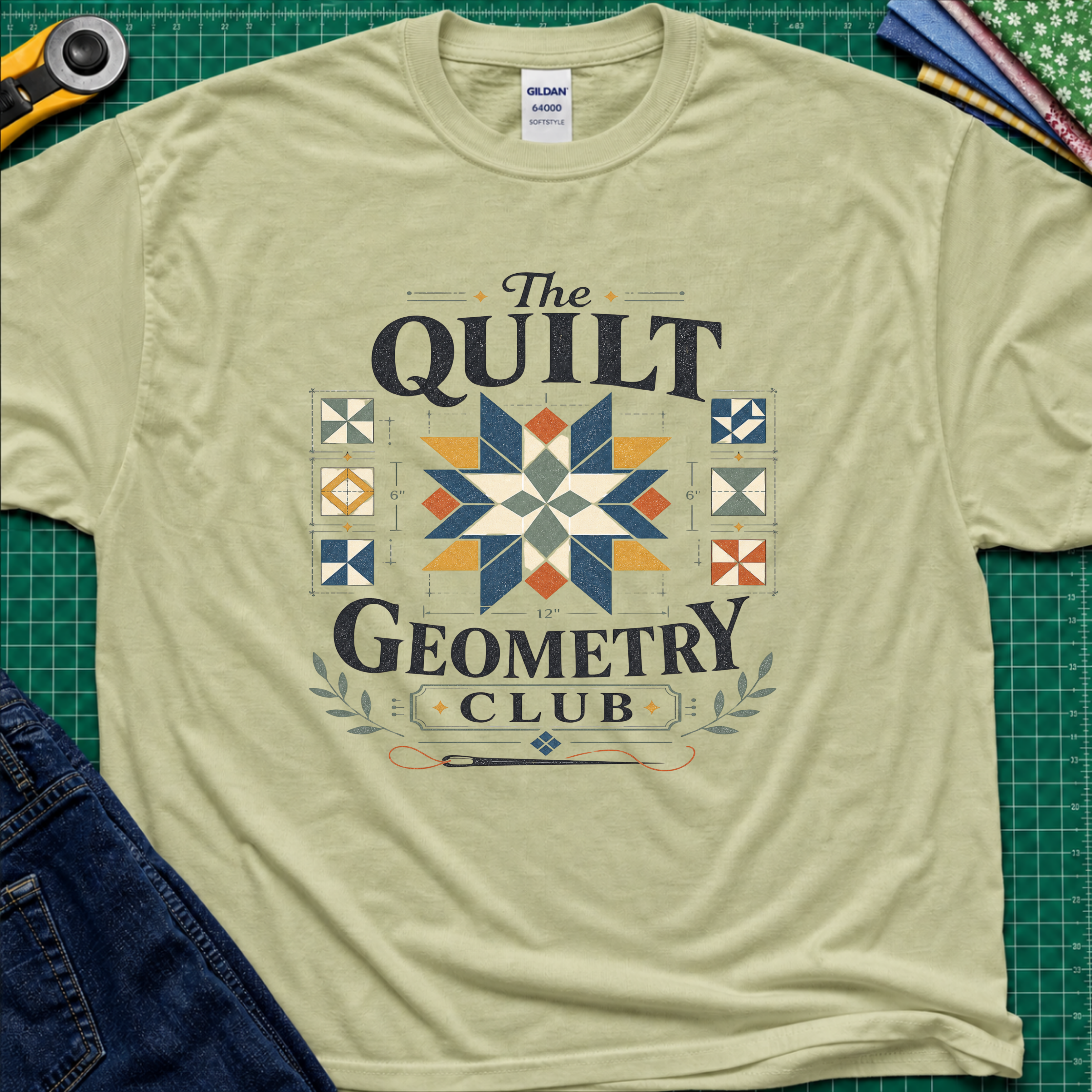 A pistachio green Gildan t-shirt with the words 'The Quilt Geometry Club' surround a colourful quilt block with smaller quilt blocks on either side. The shirt lays flat on a green cutting mat with craft tools and jeans visible around the edges.