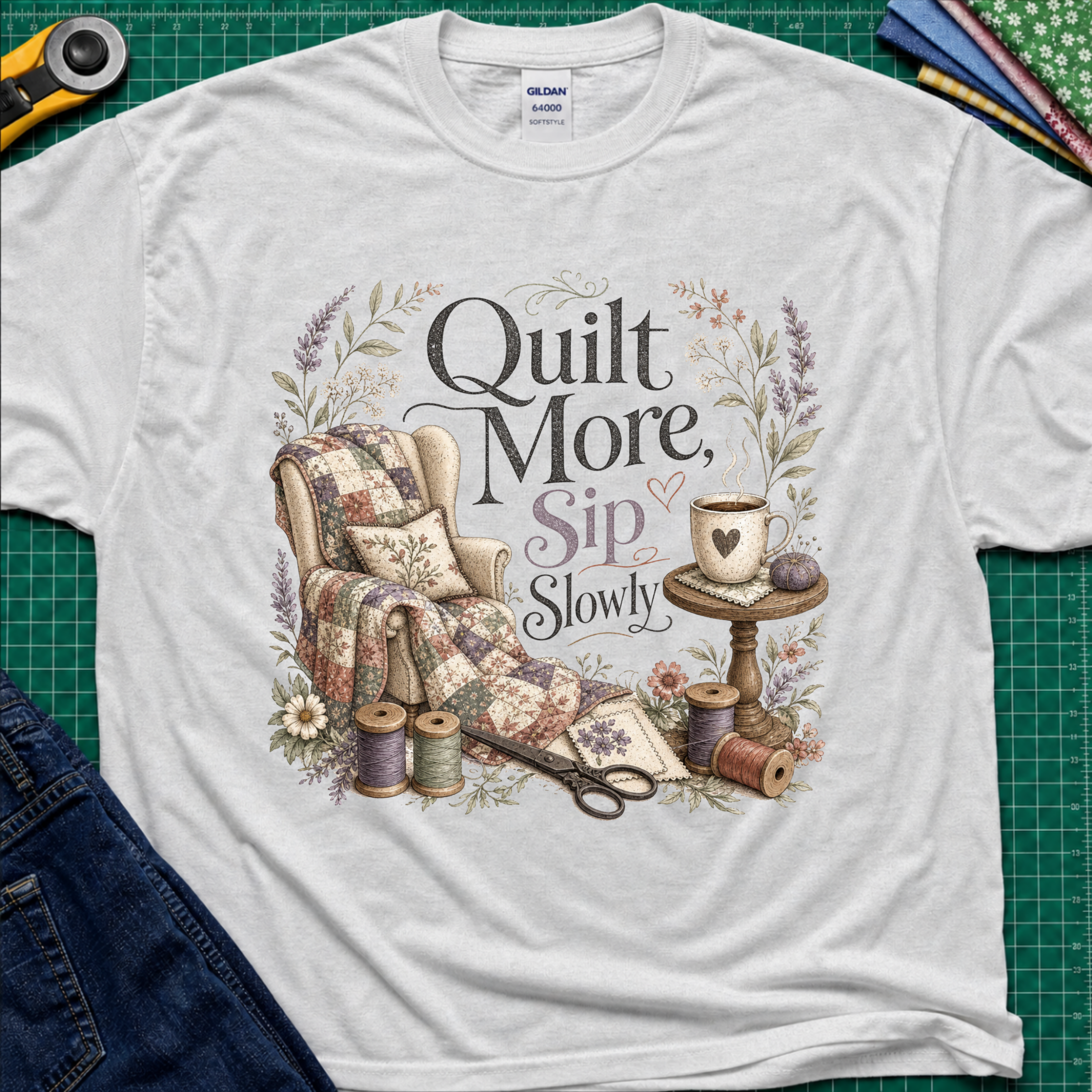 A light grey Gildan t-shirt with a vintage-style quilted graphic that reads 'Quilt More, Sip Slowly' surrounded by floral details, shown in a flat-lay arrangement on a green cutting mat with sewing props.
