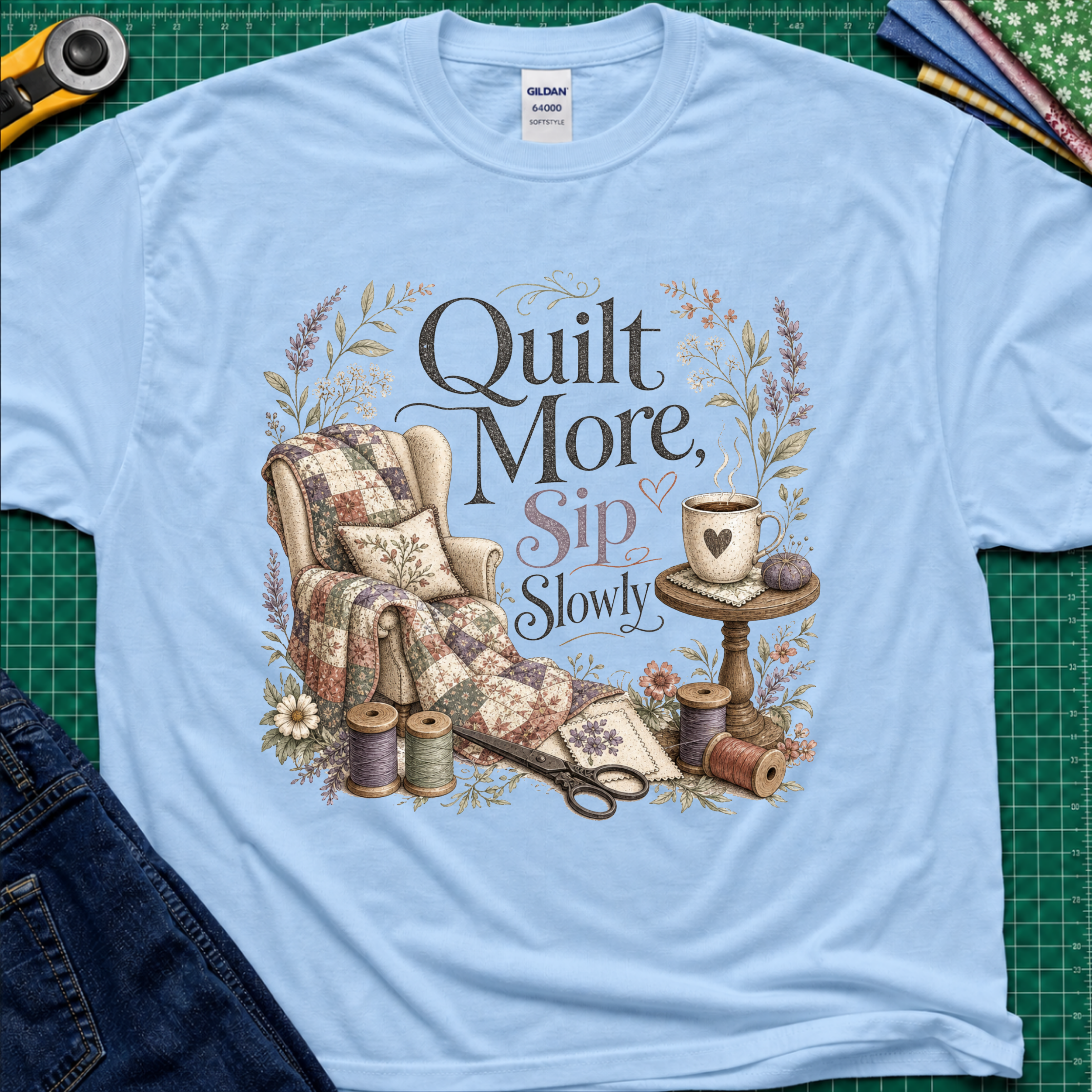 A light blue Gildan t-shirt with a vintage-style quilted graphic that reads 'Quilt More, Sip Slowly' surrounded by floral details, shown in a flat-lay arrangement on a green cutting mat with sewing props.