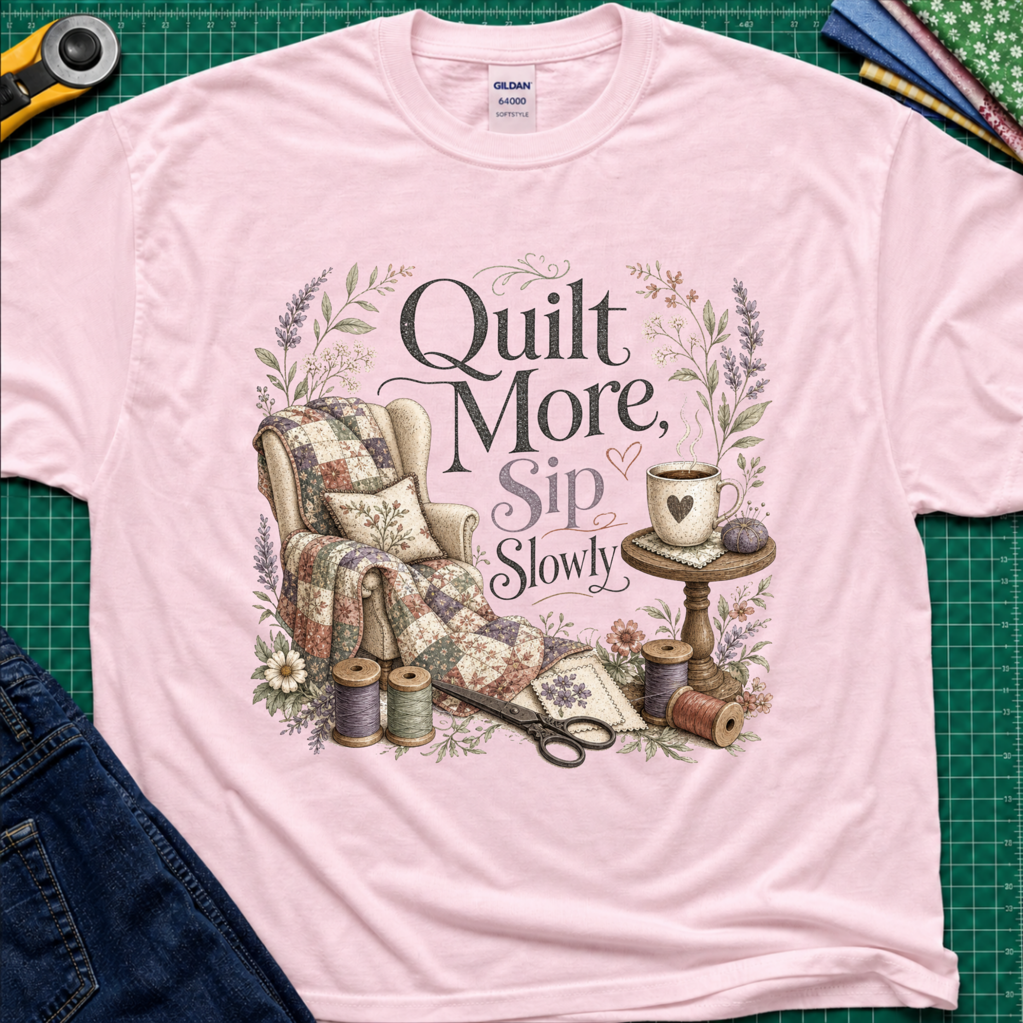 A light pink Gildan t-shirt with a vintage-style quilted graphic that reads 'Quilt More, Sip Slowly' surrounded by floral details, shown in a flat-lay arrangement on a green cutting mat with sewing props.