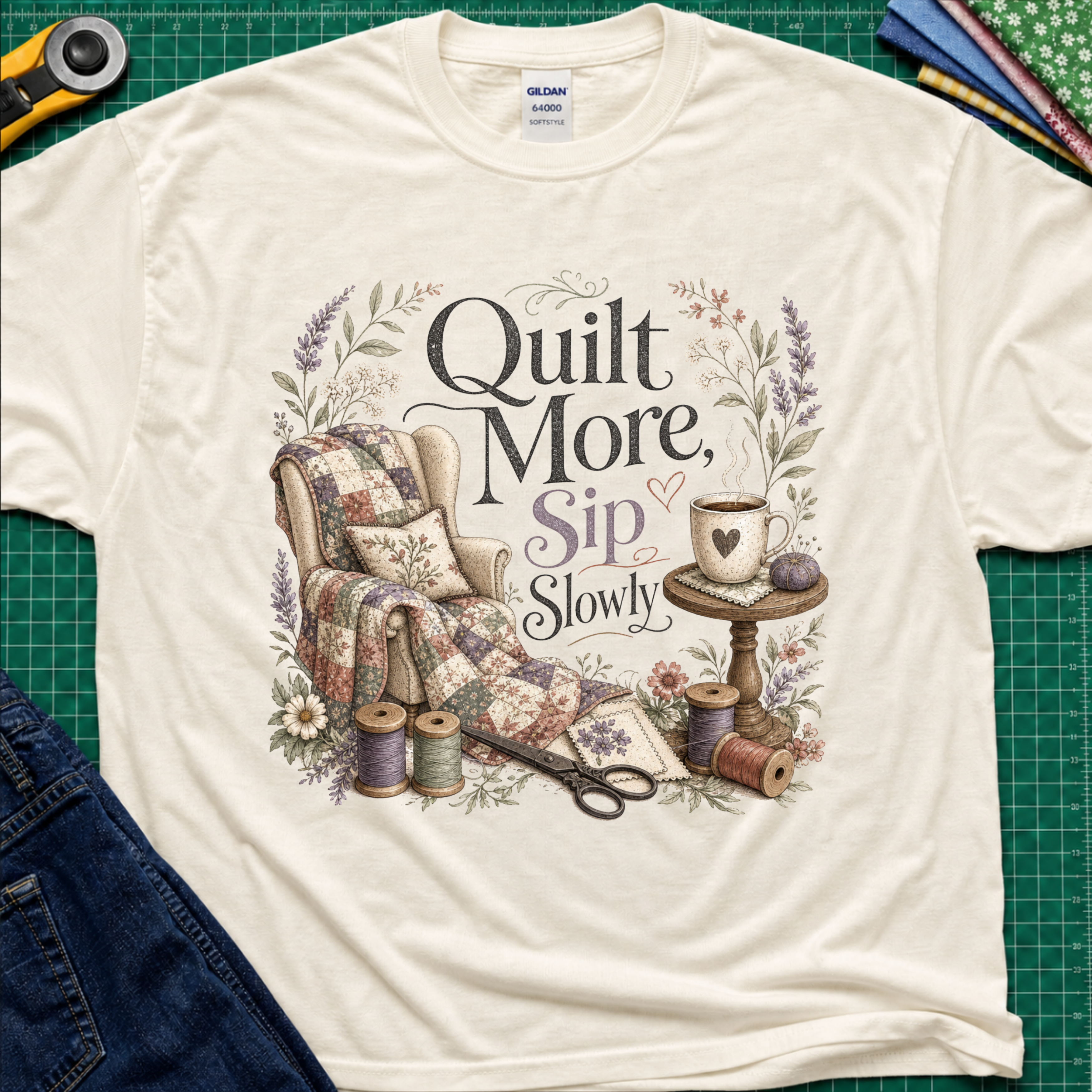 A light cream Gildan t-shirt with a vintage-style quilted graphic that reads 'Quilt More, Sip Slowly' surrounded by floral details, shown in a flat-lay arrangement on a green cutting mat with sewing props.