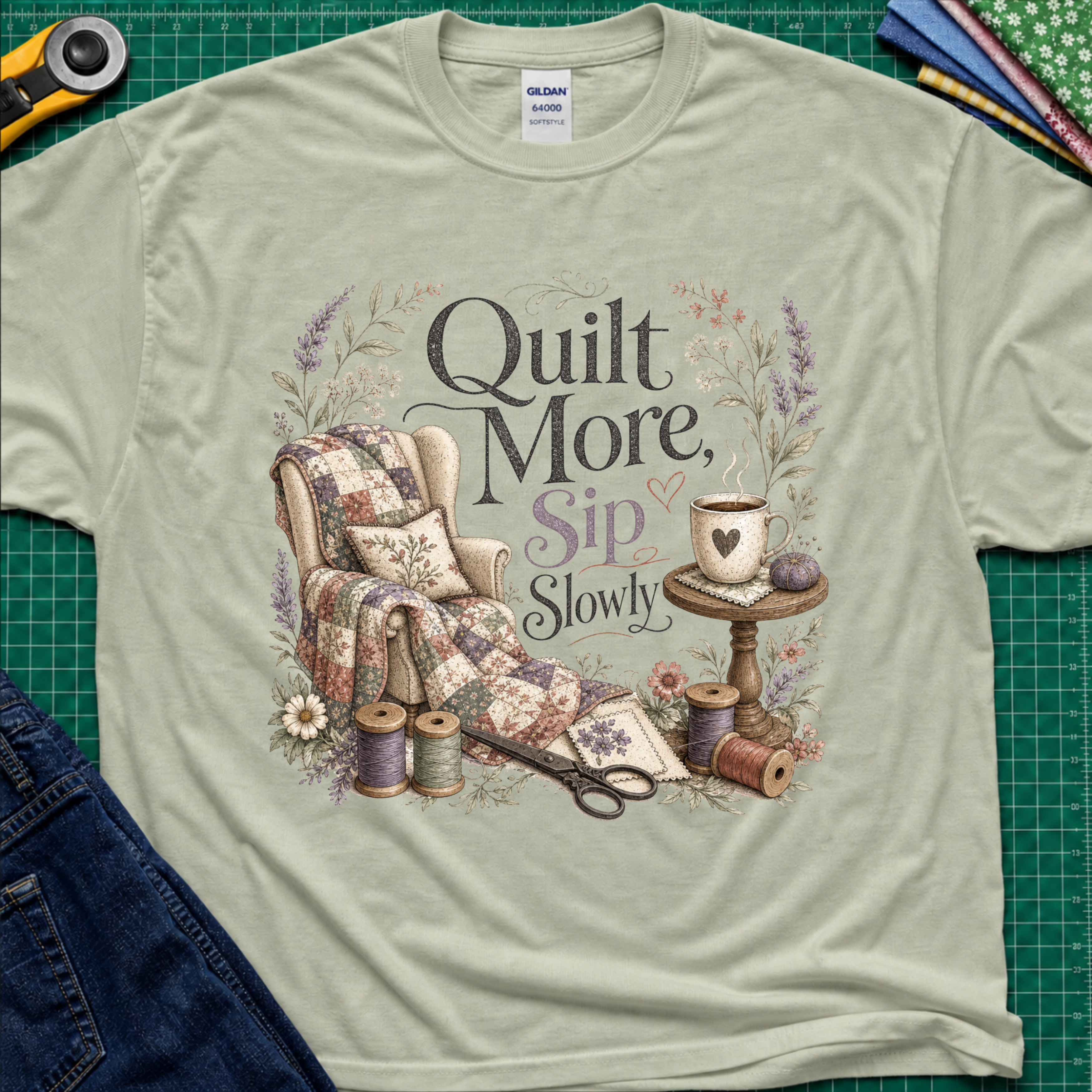 A sage green Gildan t-shirt with a vintage-style quilted graphic that reads 'Quilt More, Sip Slowly' surrounded by floral details, shown in a flat-lay arrangement on a green cutting mat with sewing props.