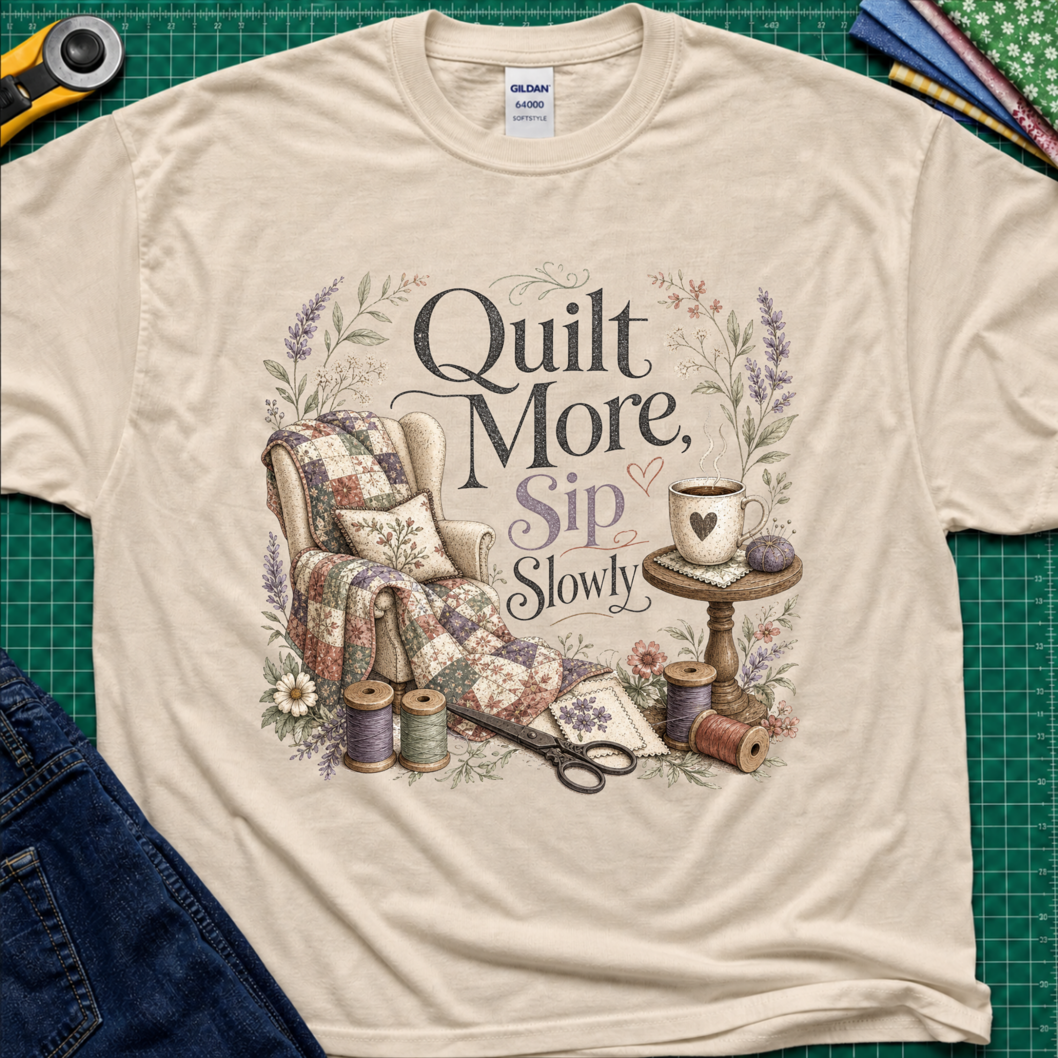 A dark cream Gildan t-shirt with a vintage-style quilted graphic that reads 'Quilt More, Sip Slowly' surrounded by floral details, shown in a flat-lay arrangement on a green cutting mat with sewing props.