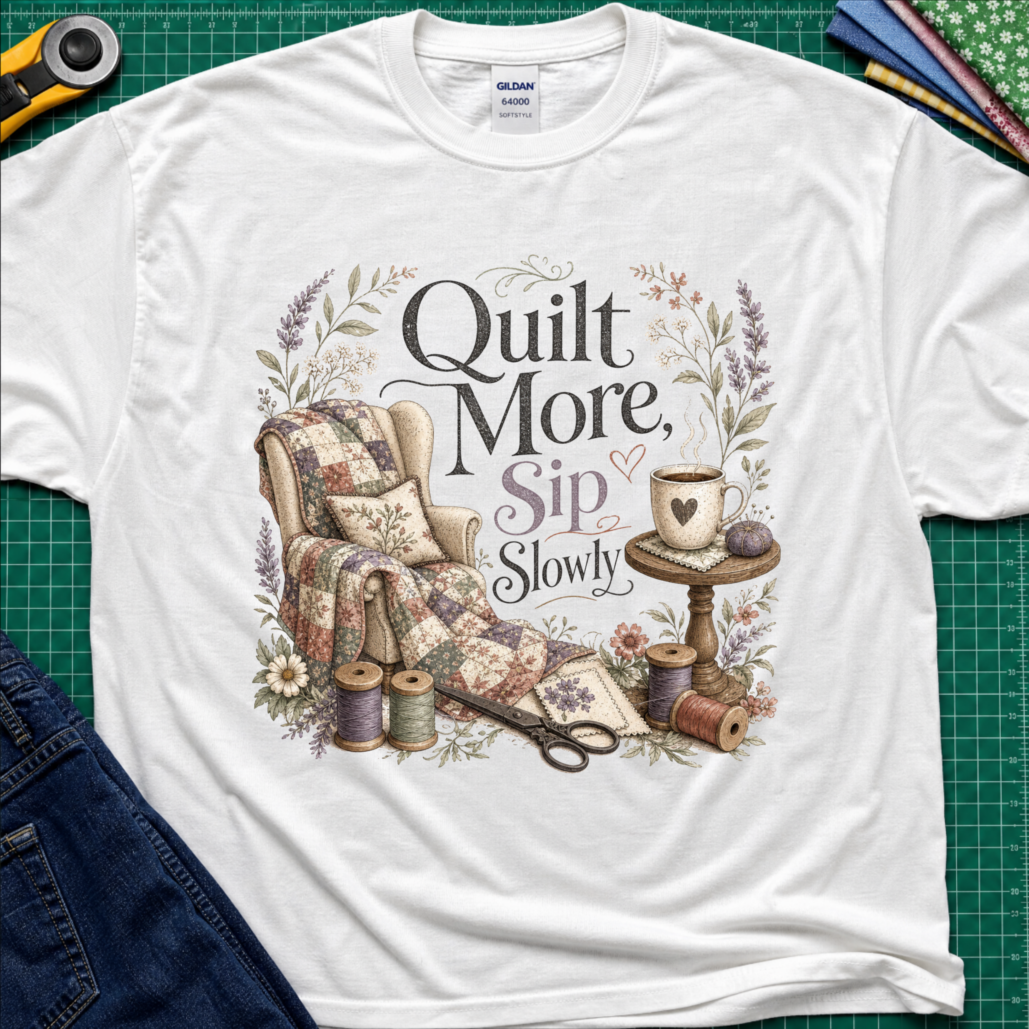 A white Gildan t-shirt with a vintage-style quilted graphic that reads 'Quilt More, Sip Slowly' surrounded by floral details, shown in a flat-lay arrangement on a green cutting mat with sewing props.