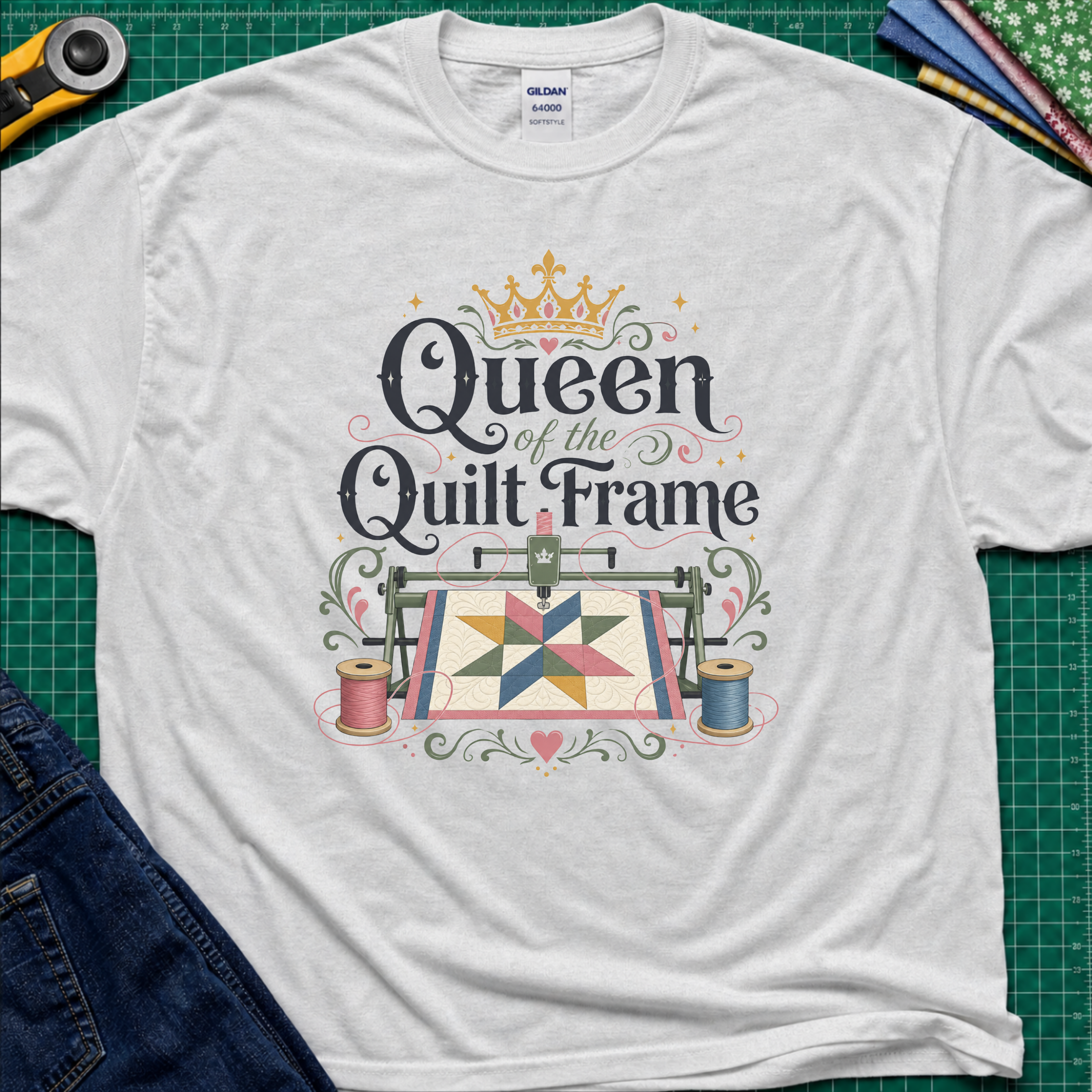 A light grey Gildan t-shirt featuring a colorful graphic that reads 'Queen of the Quilt Frame' with a crown and a quilt design, laid flat on a green cutting mat with sewing tools and jeans as props.
