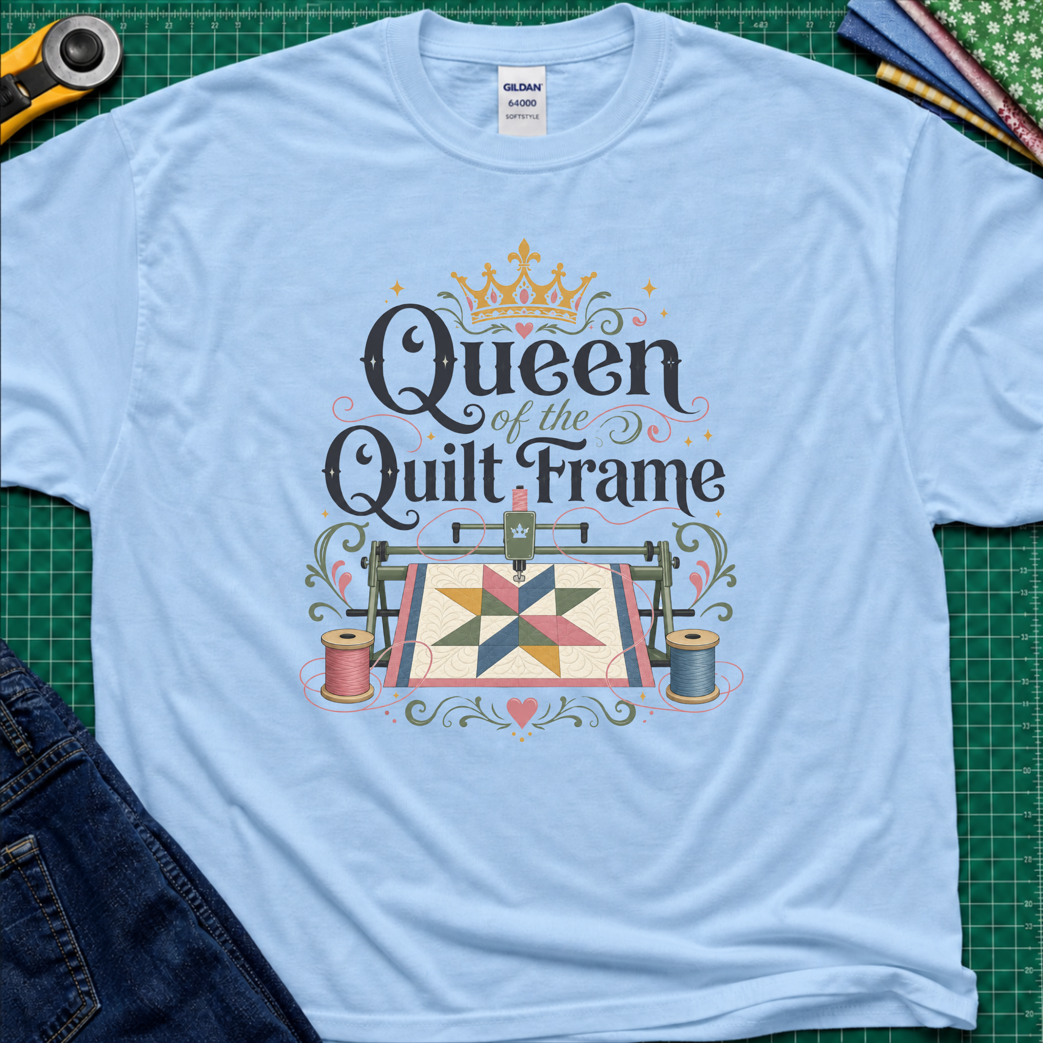 A light blue Gildan t-shirt featuring a colorful graphic that reads 'Queen of the Quilt Frame' with a crown and a quilt design, laid flat on a green cutting mat with sewing tools and jeans as props.