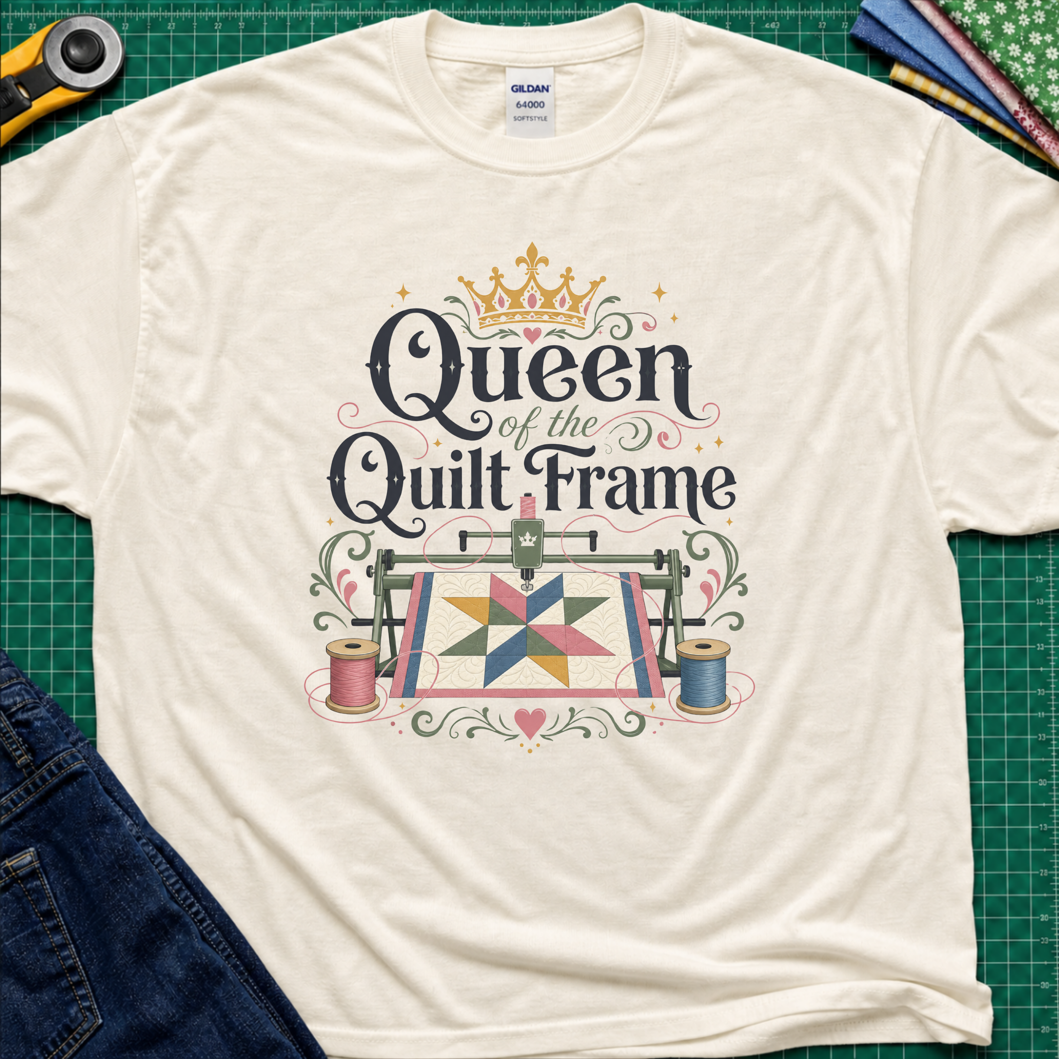 A light cream Gildan t-shirt featuring a colorful graphic that reads 'Queen of the Quilt Frame' with a crown and a quilt design, laid flat on a green cutting mat with sewing tools and jeans as props.