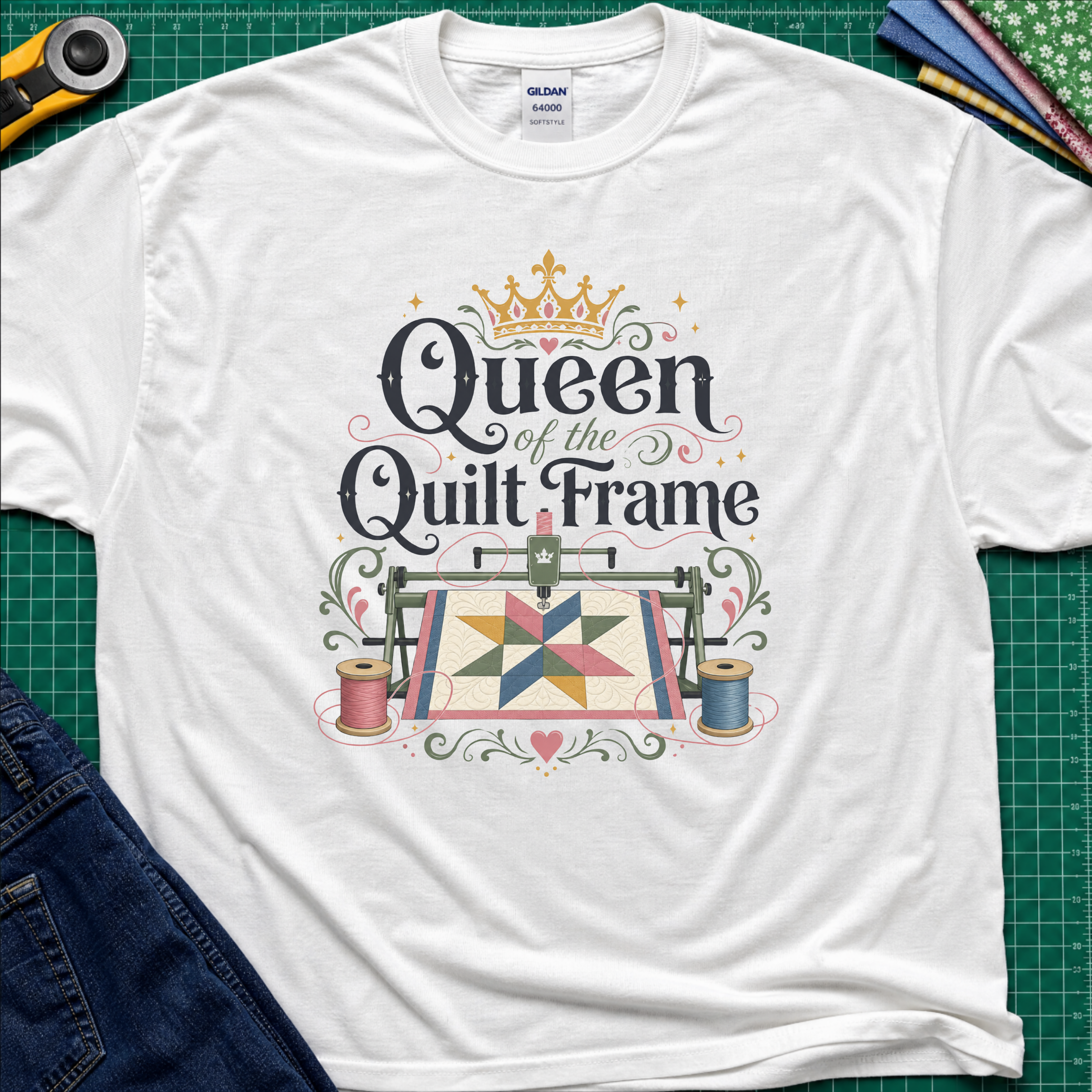 A white Gildan t-shirt featuring a colorful graphic that reads 'Queen of the Quilt Frame' with a crown and a quilt design, laid flat on a green cutting mat with sewing tools and jeans as props.