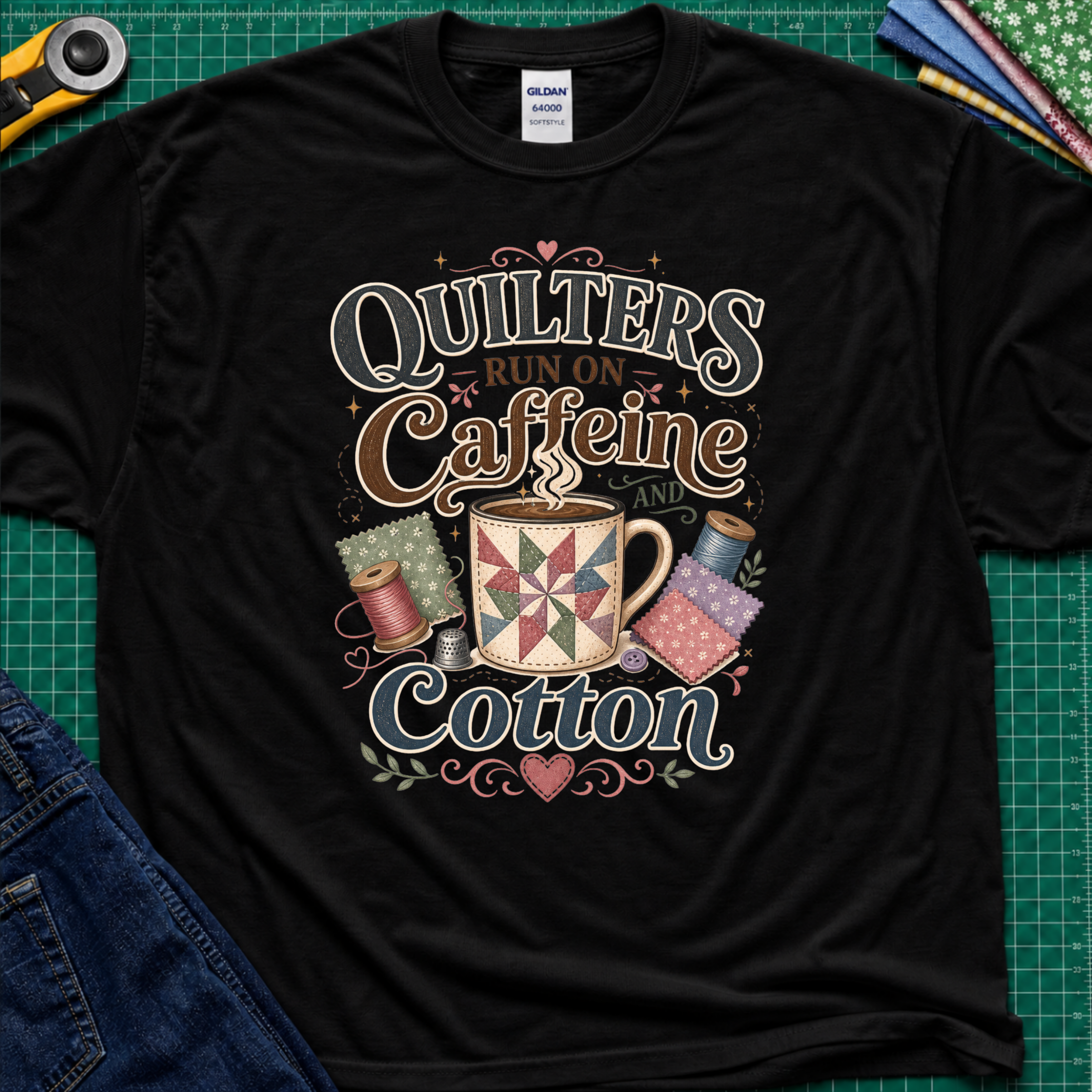 A black Gildan t-shirt with a colorful graphic that reads 'Quilters Run on Caffeine and Cotton' around an illustration of a quilted mug and spools of thread. The shirt lays flat on a green cutting mat with a rotary cutter, fabric stack and jeans partially visible in the corners.