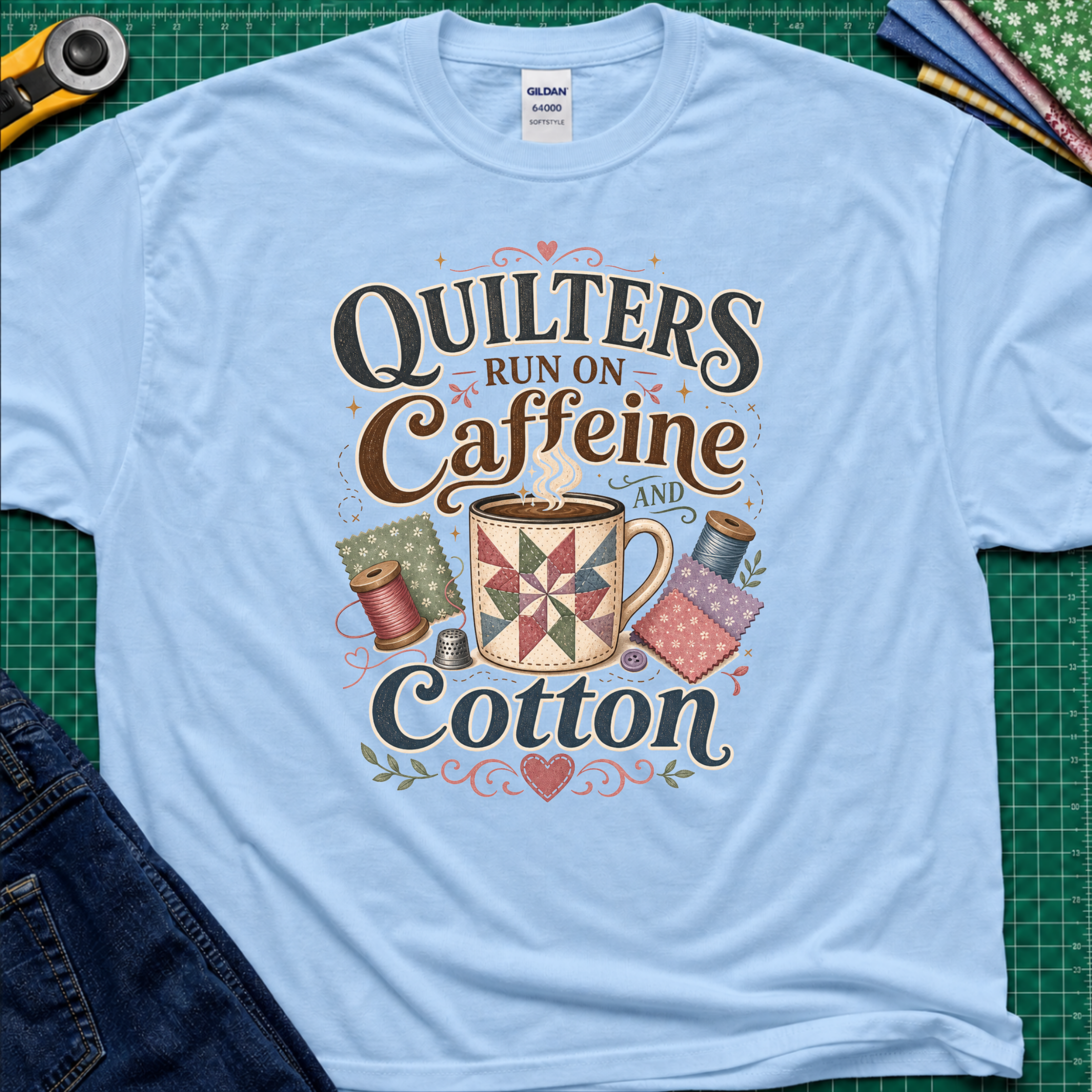 A light blue Gildan t-shirt with a colorful graphic that reads 'Quilters Run on Caffeine and Cotton' around an illustration of a quilted mug and spools of thread. The shirt lays flat on a green cutting mat with a rotary cutter, fabric stack and jeans partially visible in the corners.