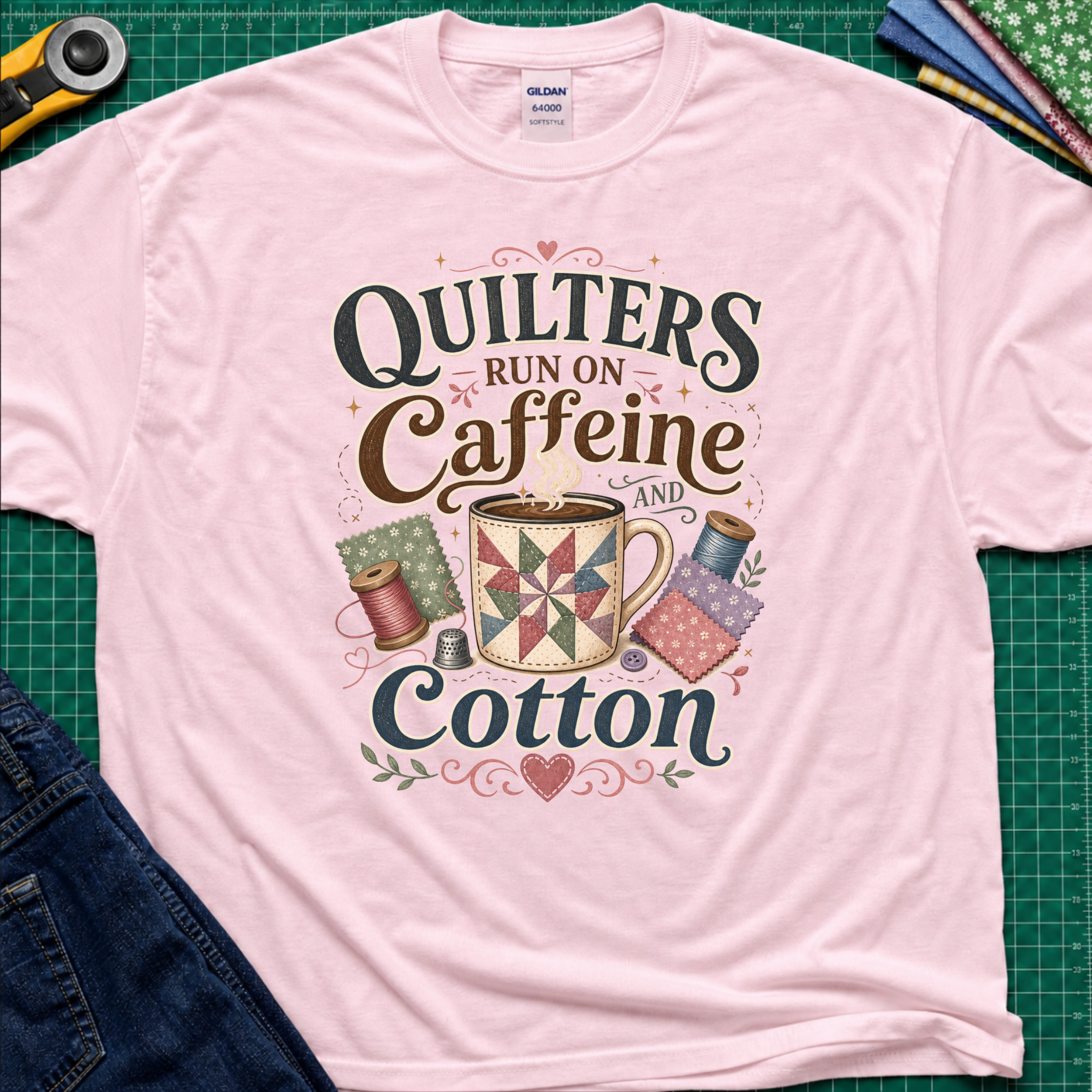 A light pink Gildan t-shirt with a colorful graphic that reads 'Quilters Run on Caffeine and Cotton' around an illustration of a quilted mug and spools of thread. The shirt lays flat on a green cutting mat with a rotary cutter, fabric stack and jeans partially visible in the corners.