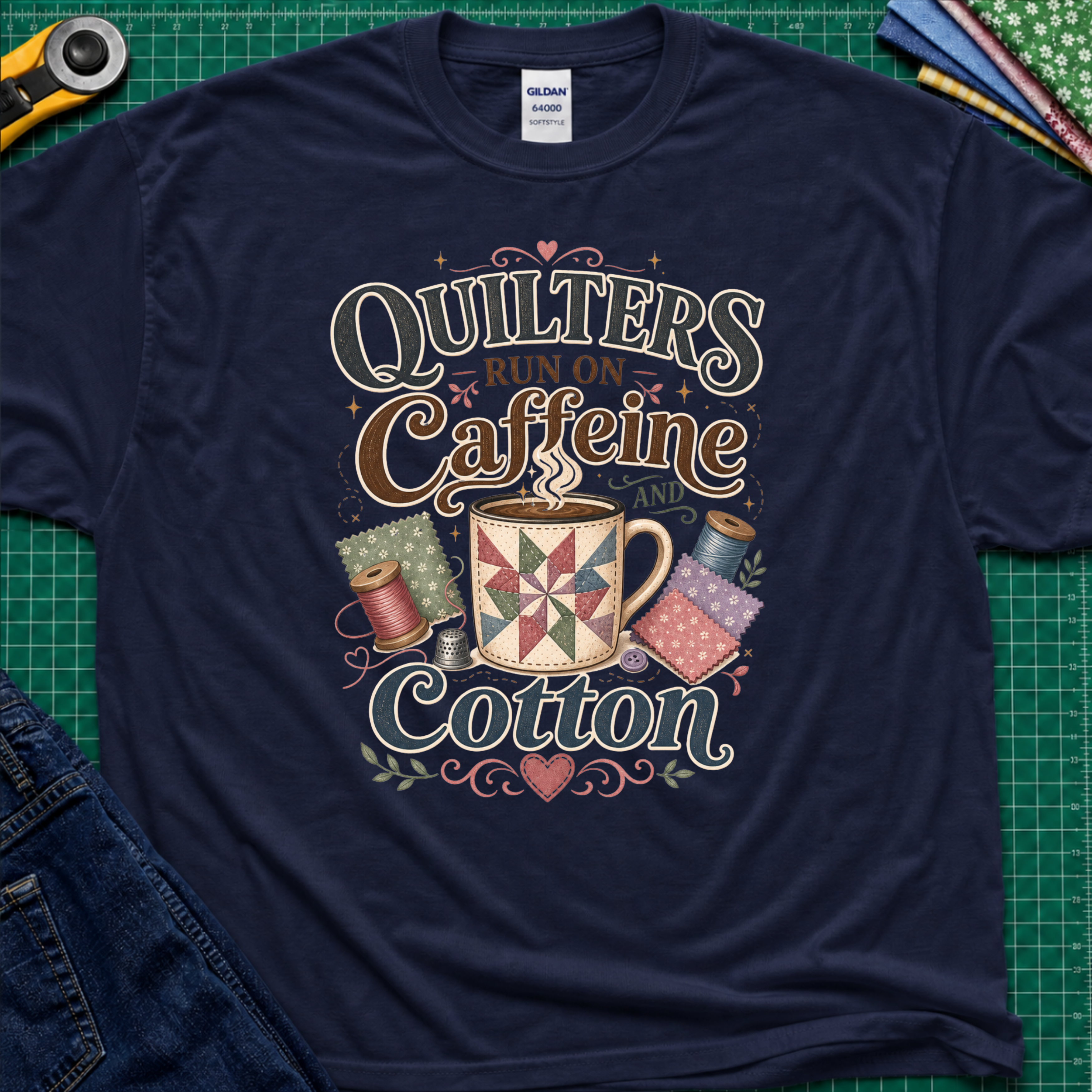 A navy blue Gildan t-shirt with a colorful graphic that reads 'Quilters Run on Caffeine and Cotton' around an illustration of a quilted mug and spools of thread. The shirt lays flat on a green cutting mat with a rotary cutter, fabric stack and jeans partially visible in the corners.