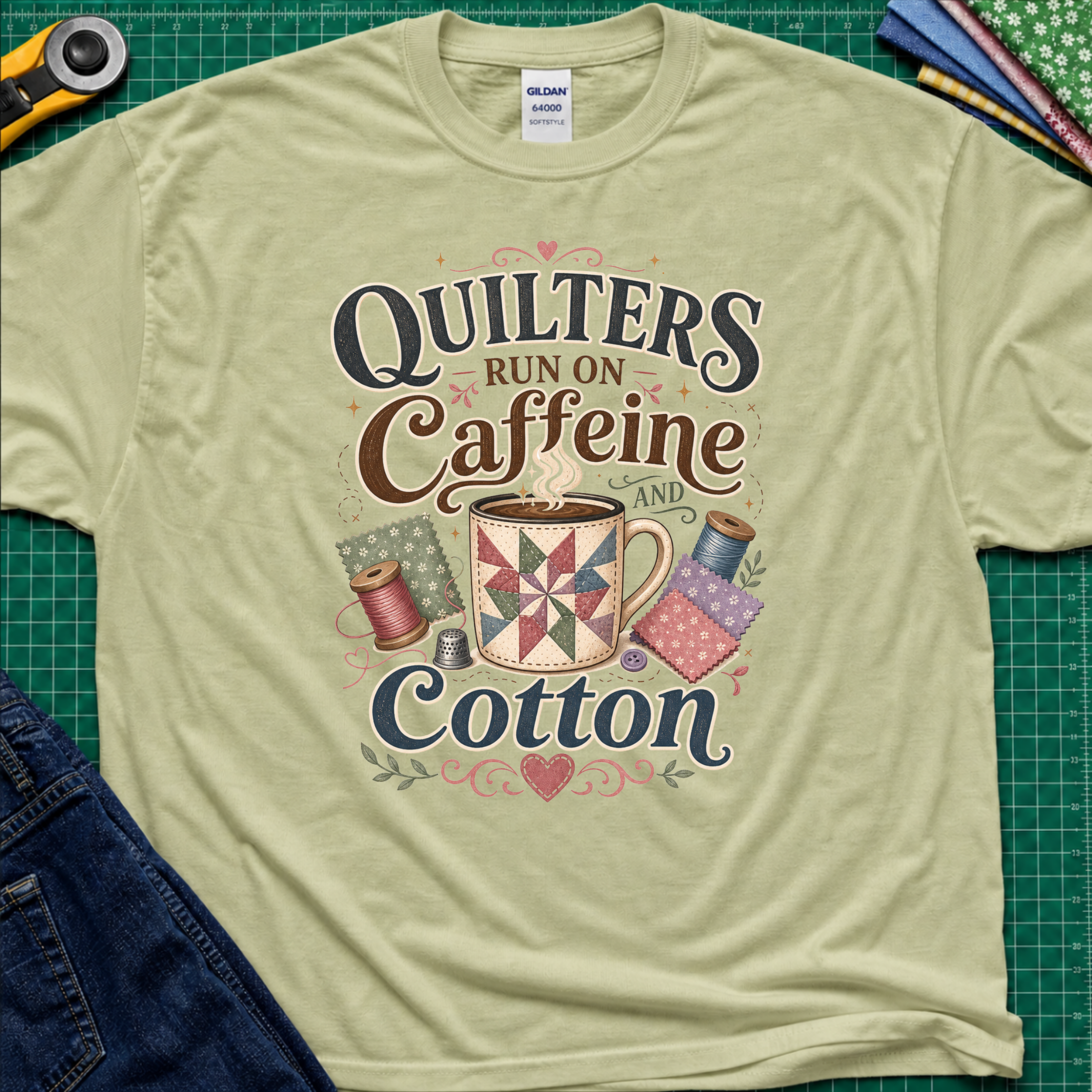 A pistachio green Gildan t-shirt with a colorful graphic that reads 'Quilters Run on Caffeine and Cotton' around an illustration of a quilted mug and spools of thread. The shirt lays flat on a green cutting mat with a rotary cutter, fabric stack and jeans partially visible in the corners.