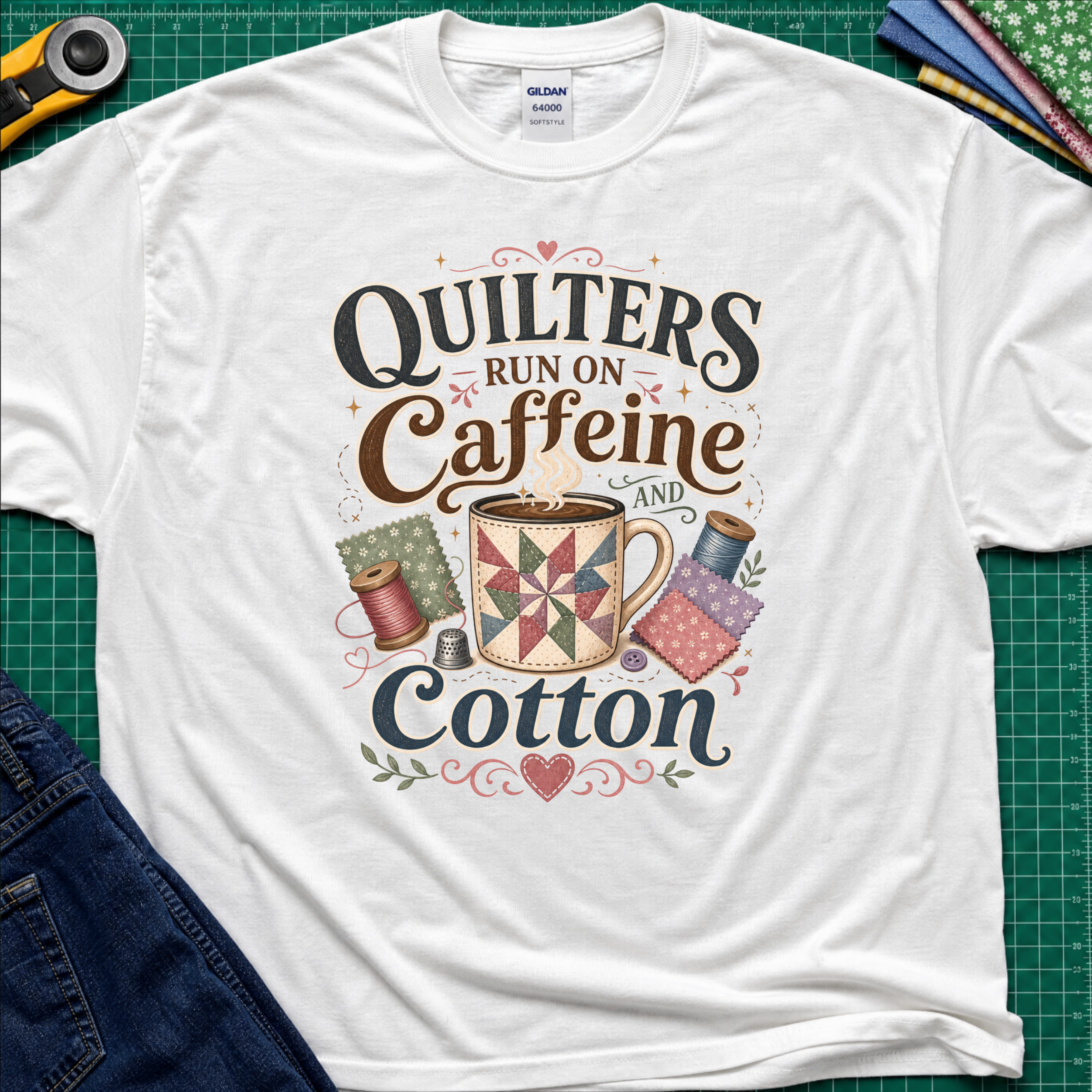 A white Gildan t-shirt with a colorful graphic that reads 'Quilters Run on Caffeine and Cotton' around an illustration of a quilted mug and spools of thread. The shirt lays flat on a green cutting mat with a rotary cutter, fabric stack and jeans partially visible in the corners.