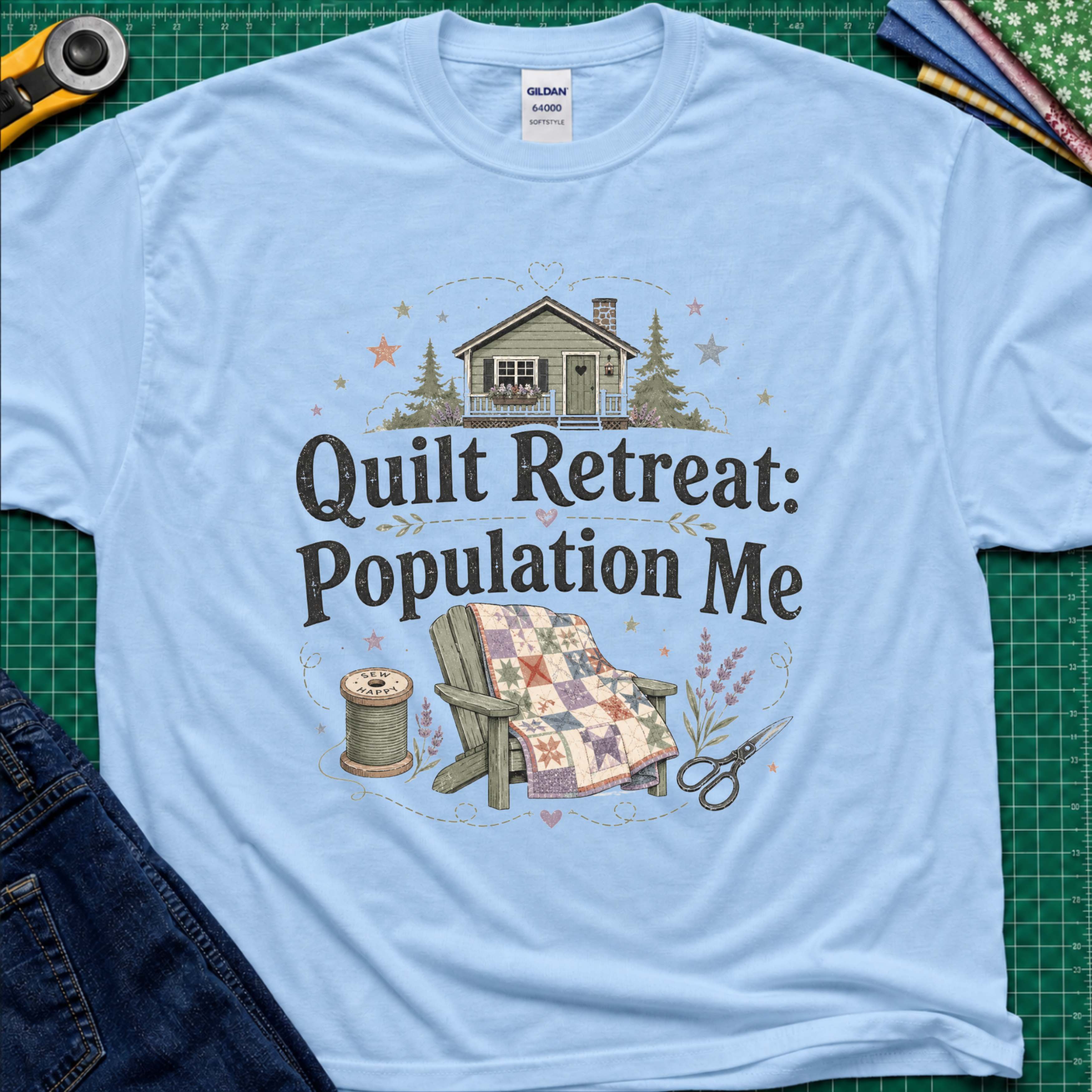A light blue Gildan t-shirt with a 'Quilt Retreat: Population Me' graphic print, styled in a flat-lay on a green cutting mat with sewing props.