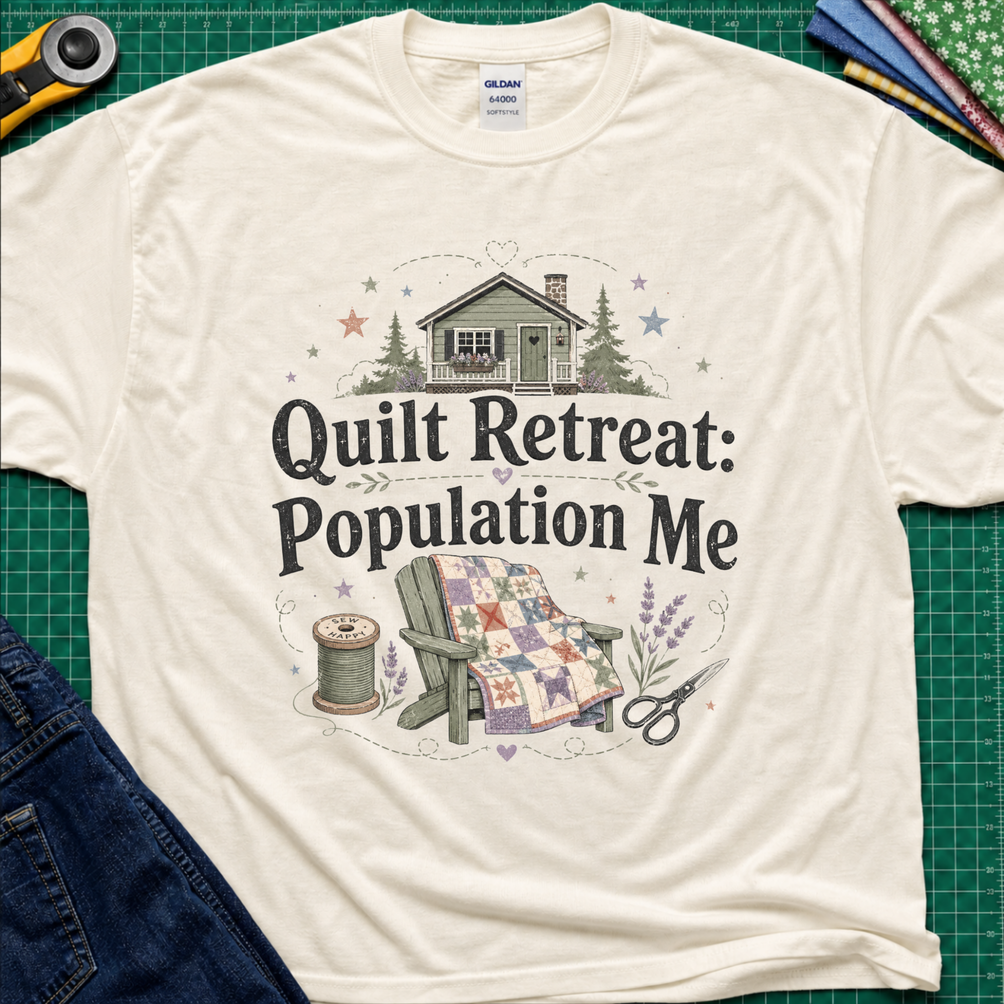A light cream Gildan t-shirt with a 'Quilt Retreat: Population Me' graphic print, styled in a flat-lay on a green cutting mat with sewing props.