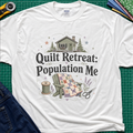 A white Gildan t-shirt with a 'Quilt Retreat: Population Me' graphic print, styled in a flat-lay on a green cutting mat with sewing props.