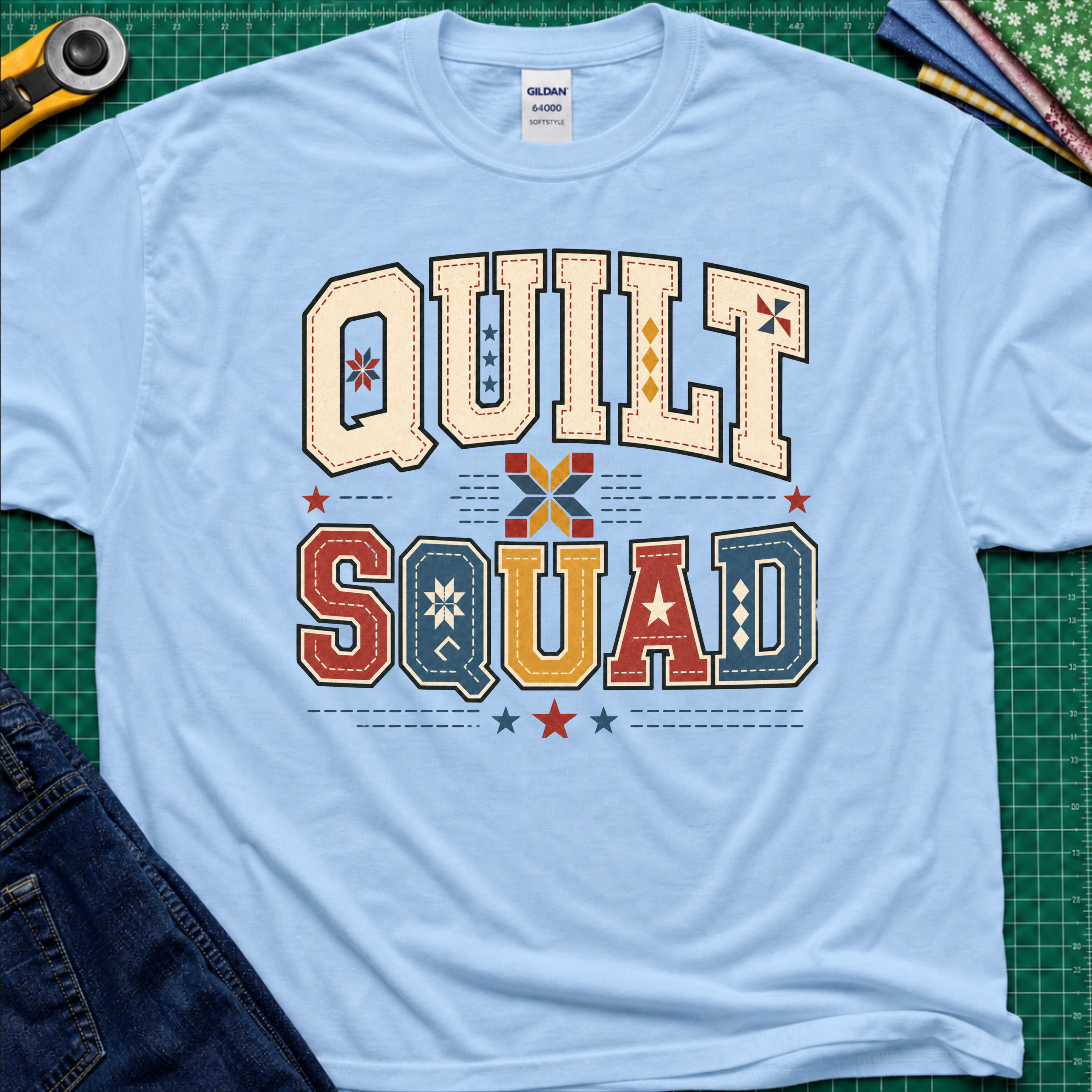 A light blue Gildan t-shirt featuring a colorful retro graphic that reads 'QUILT SQUAD' in patchwork-style letters, laid flat on a green cutting mat with craft tools and denim partially visible around the edges.