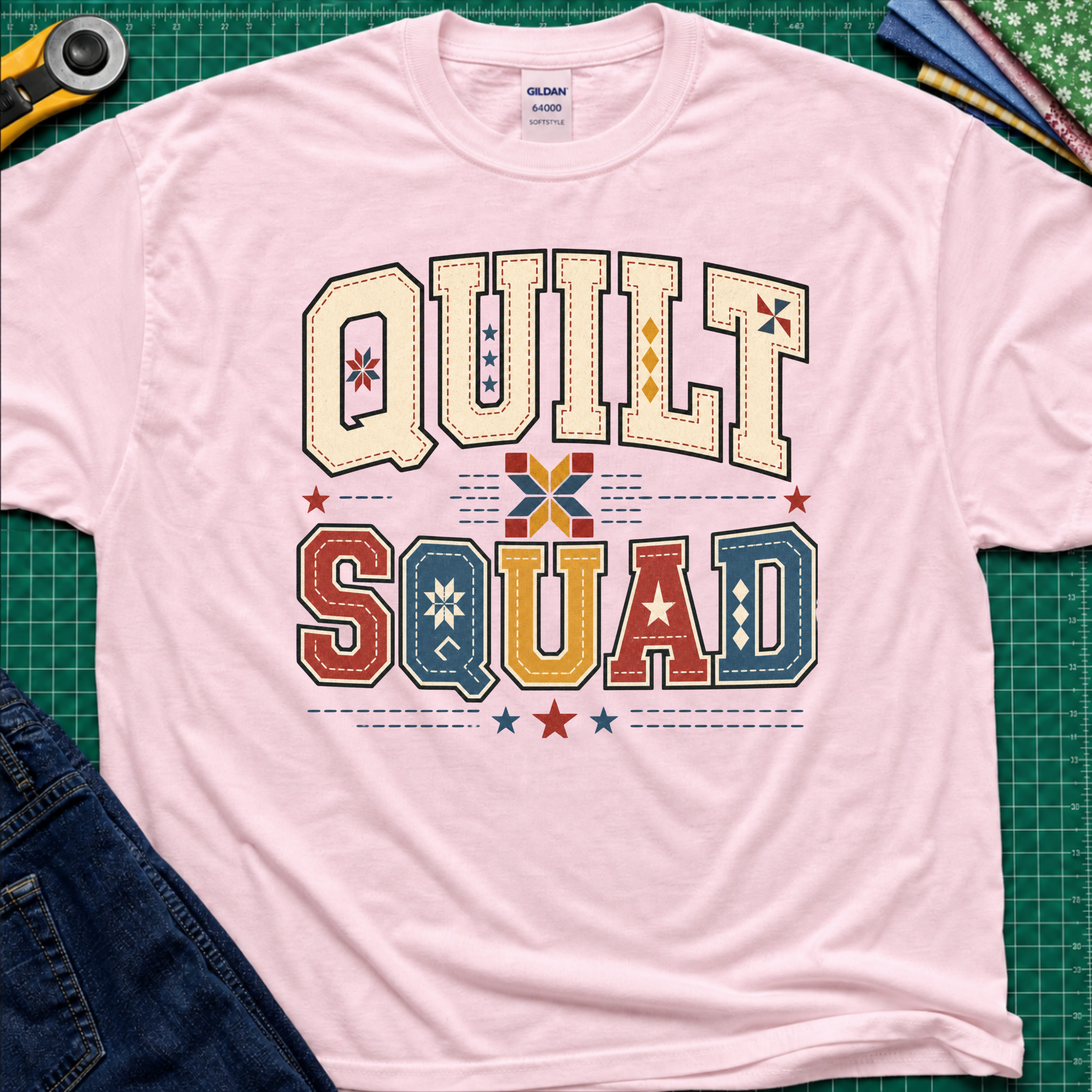 A light pink Gildan t-shirt featuring a colorful retro graphic that reads 'QUILT SQUAD' in patchwork-style letters, laid flat on a green cutting mat with craft tools and denim partially visible around the edges.