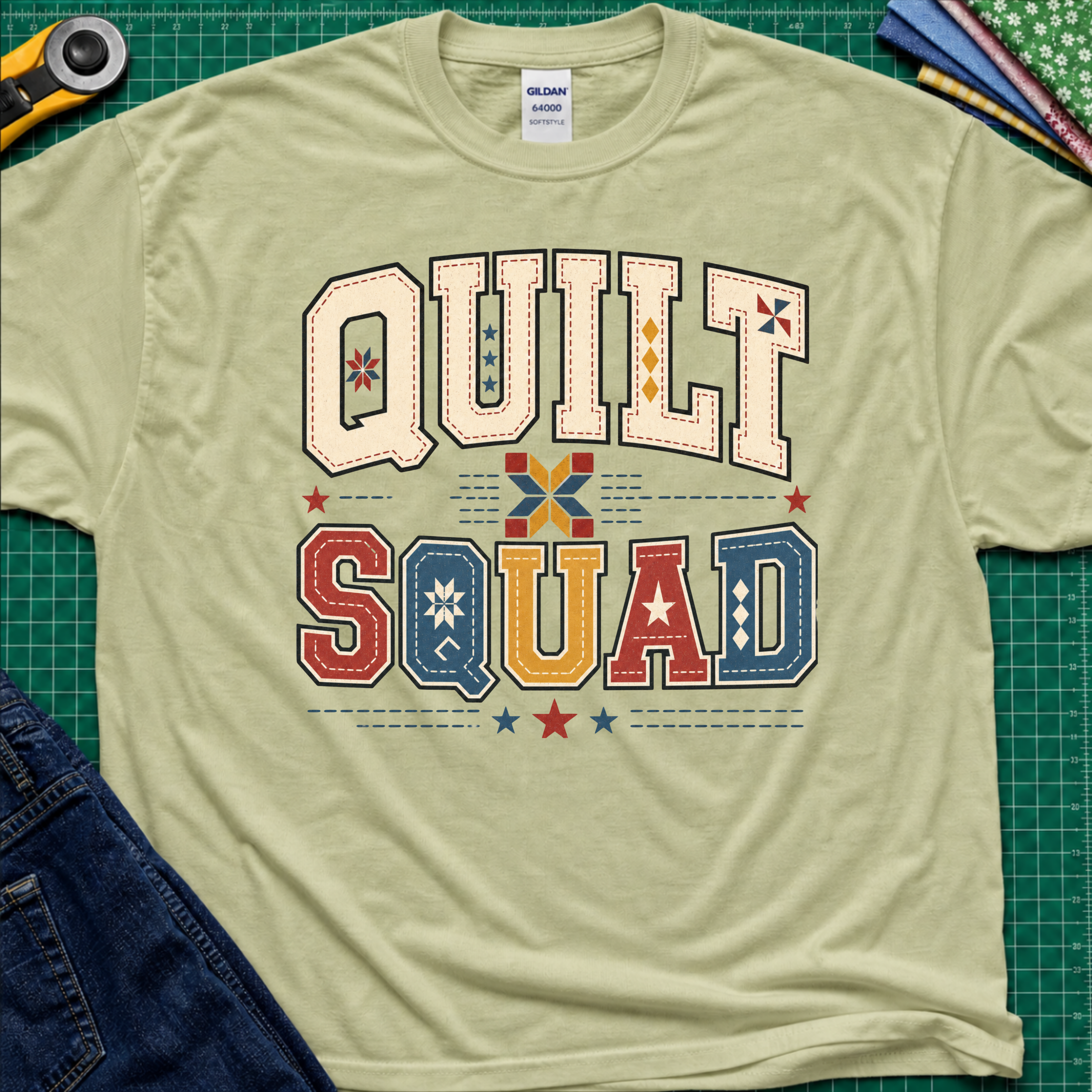 A light pistachio green Gildan t-shirt featuring a colorful retro graphic that reads 'QUILT SQUAD' in patchwork-style letters, laid flat on a green cutting mat with craft tools and denim partially visible around the edges.