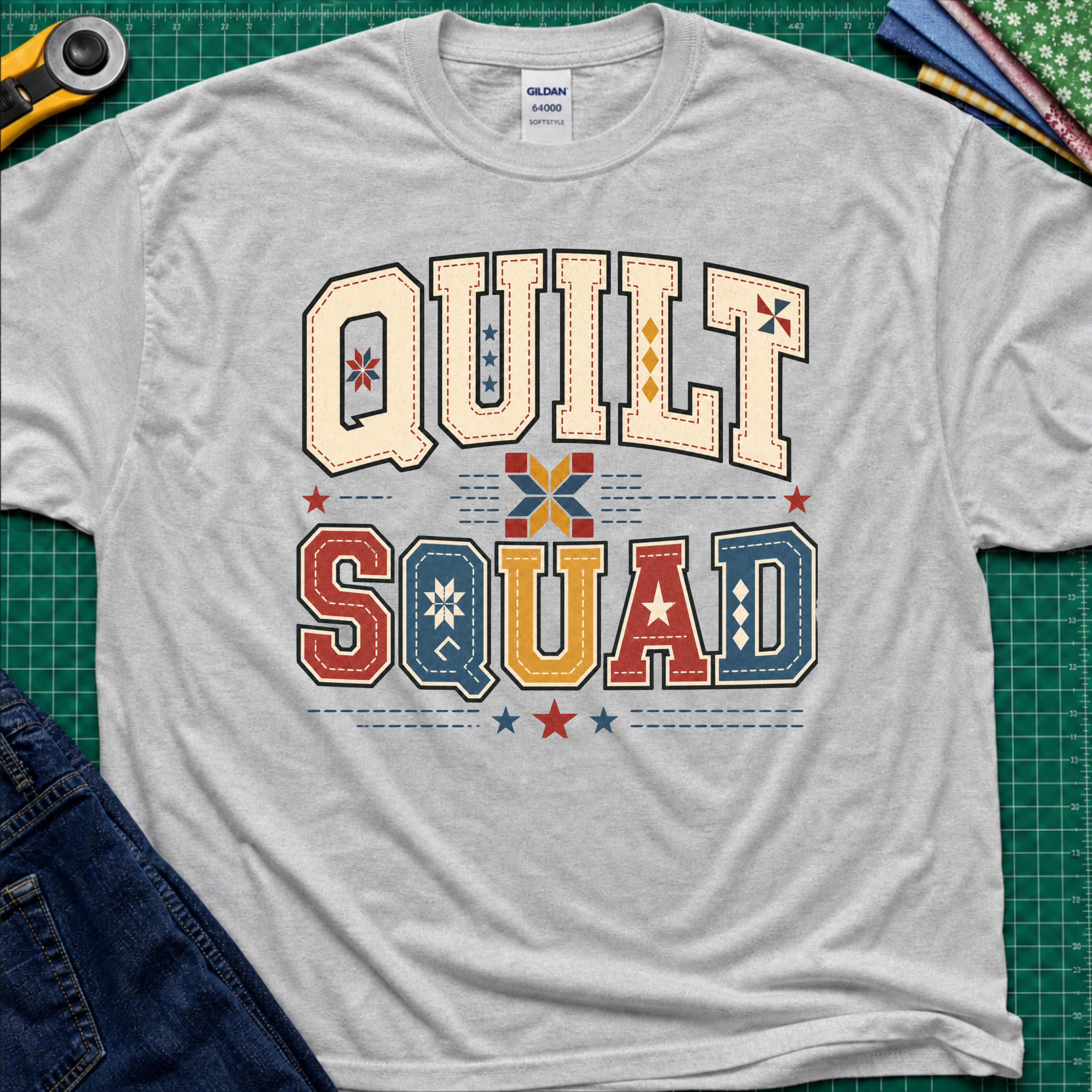 A light sports grey Gildan t-shirt featuring a colorful retro graphic that reads 'QUILT SQUAD' in patchwork-style letters, laid flat on a green cutting mat with craft tools and denim partially visible around the edges.