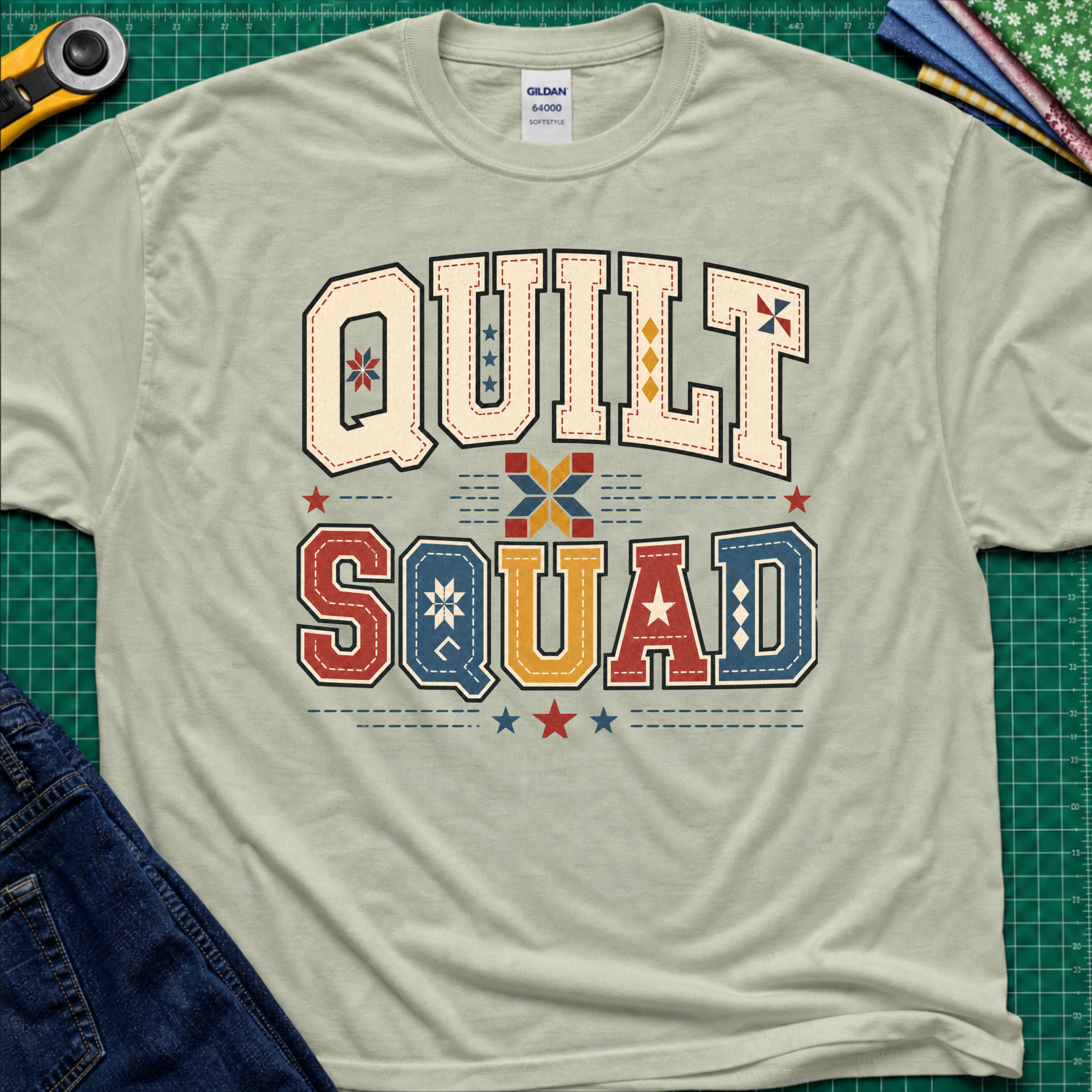 A light sage green Gildan t-shirt featuring a colorful retro graphic that reads 'QUILT SQUAD' in patchwork-style letters, laid flat on a green cutting mat with craft tools and denim partially visible around the edges.