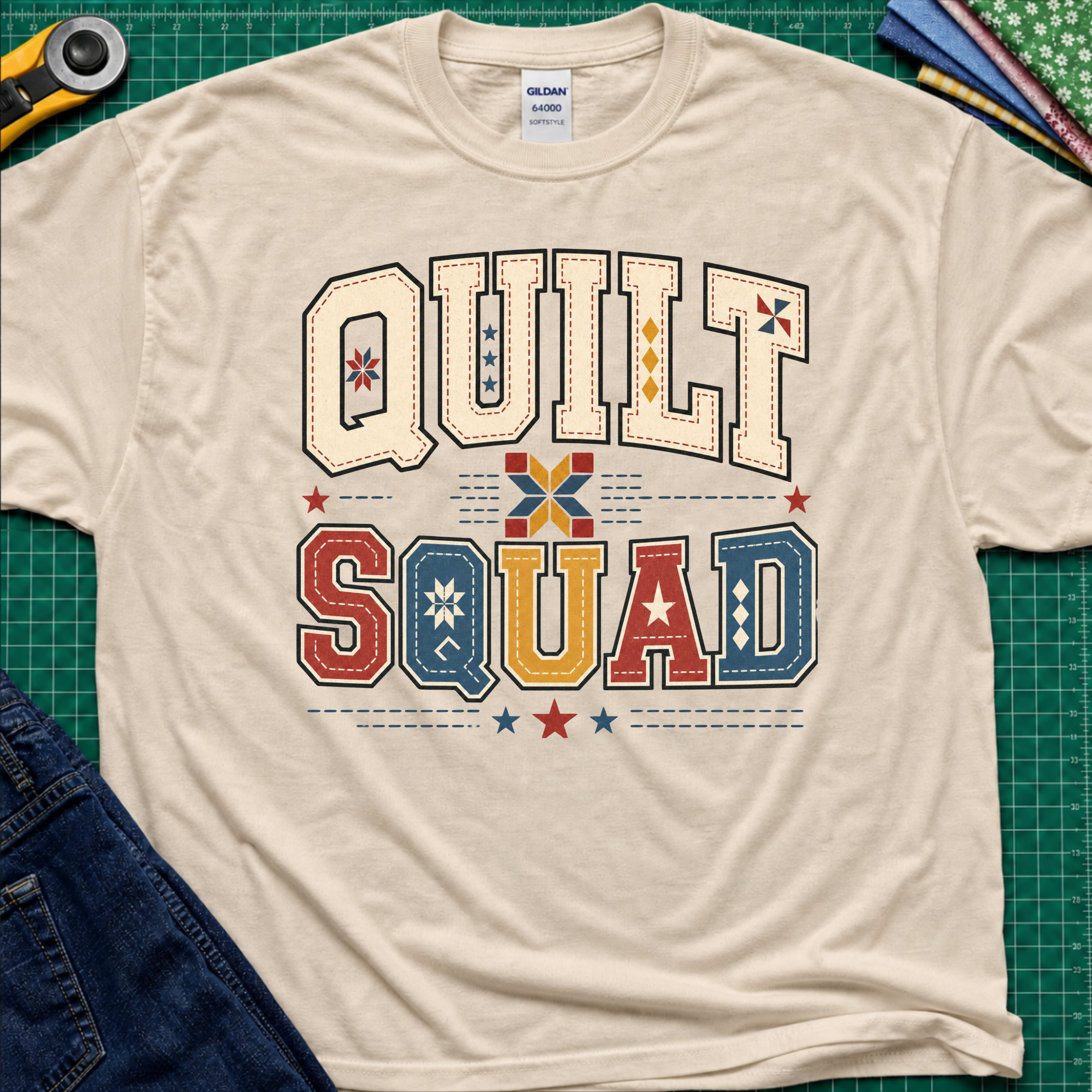 A dark cream Gildan t-shirt featuring a colorful retro graphic that reads 'QUILT SQUAD' in patchwork-style letters, laid flat on a green cutting mat with craft tools and denim partially visible around the edges.