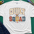 A white Gildan t-shirt featuring a colorful retro graphic that reads 'QUILT SQUAD' in patchwork-style letters, laid flat on a green cutting mat with craft tools and denim partially visible around the edges.
