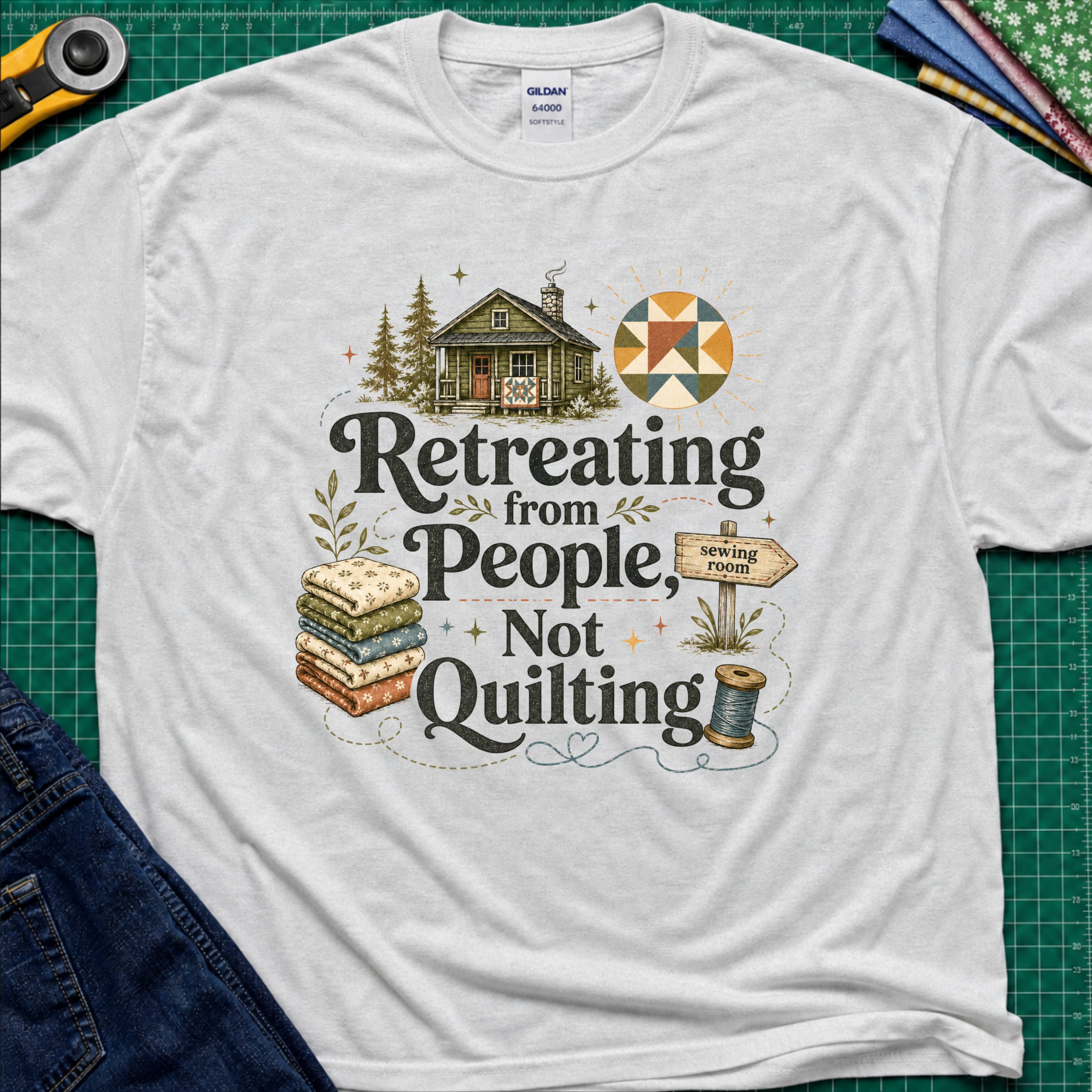 A light grey Gildan t-shirt reading 'Retreating from People Not Quilting' surrounded by illustrations of a cabin, quilts, a sewing machine, and a sign. The shirt is displayed flat on a green cutting mat with quilting tools and jeans partially visible.