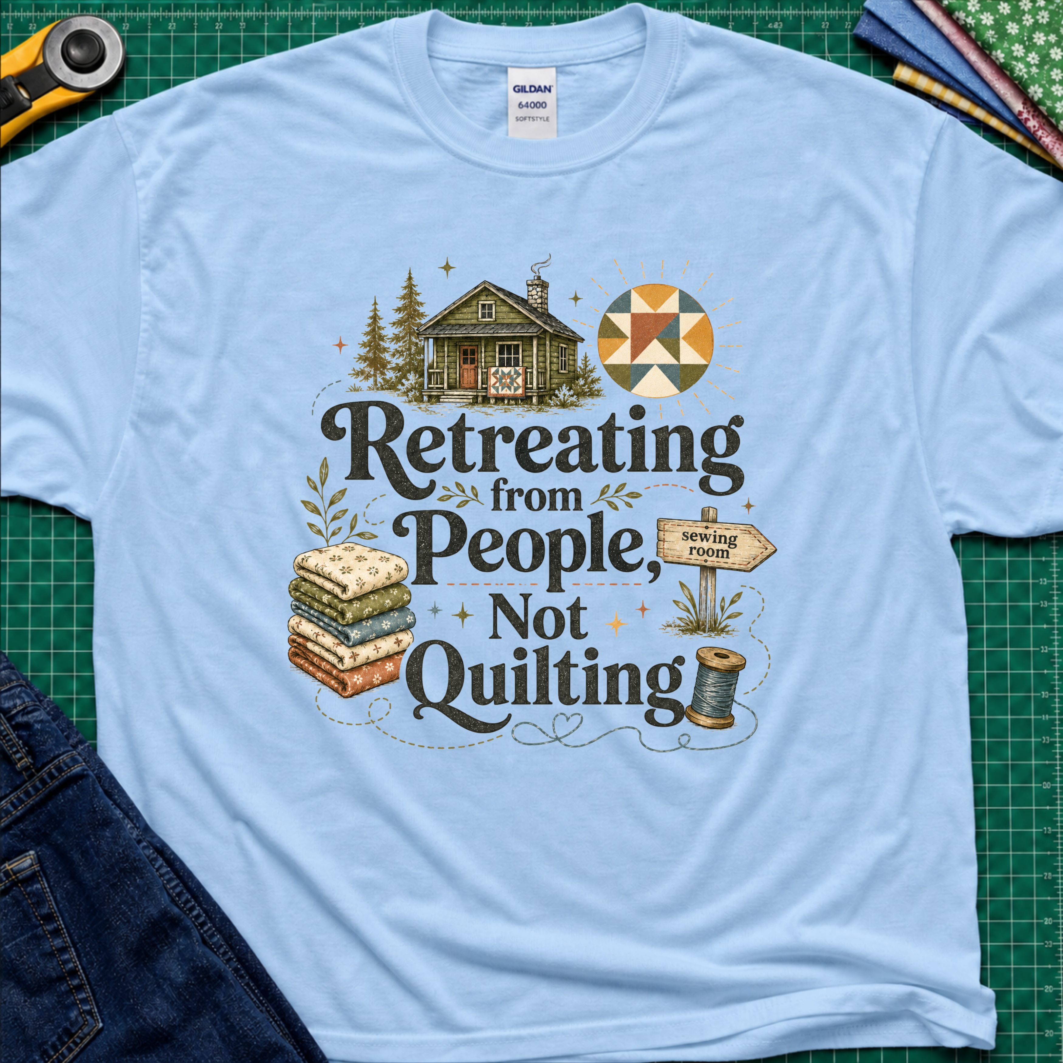 A light blue Gildan t-shirt reading 'Retreating from People Not Quilting' surrounded by illustrations of a cabin, quilts, a sewing machine, and a sign. The shirt is displayed flat on a green cutting mat with quilting tools and jeans partially visible.