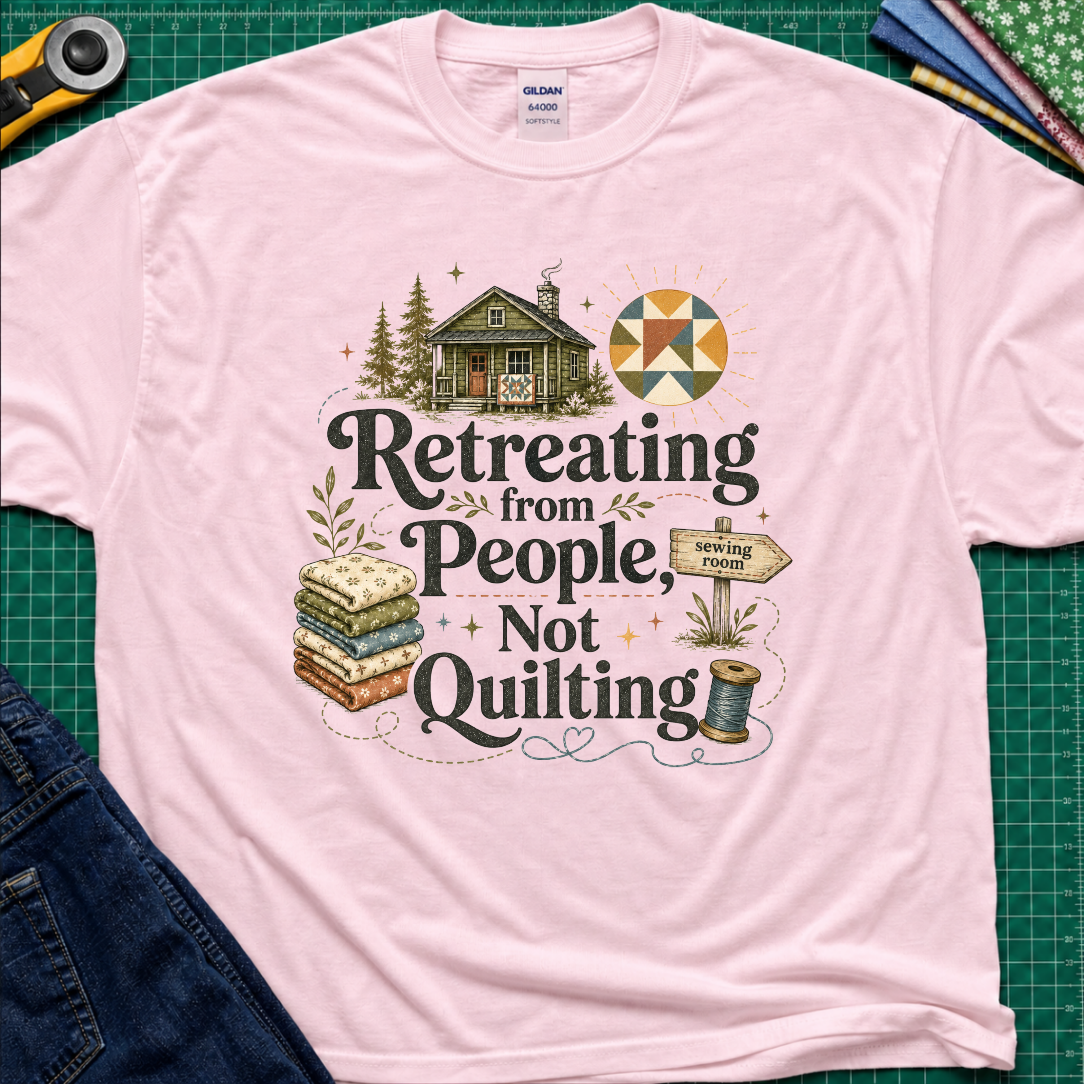 A light pink Gildan t-shirt reading 'Retreating from People Not Quilting' surrounded by illustrations of a cabin, quilts, a sewing machine, and a sign. The shirt is displayed flat on a green cutting mat with quilting tools and jeans partially visible.