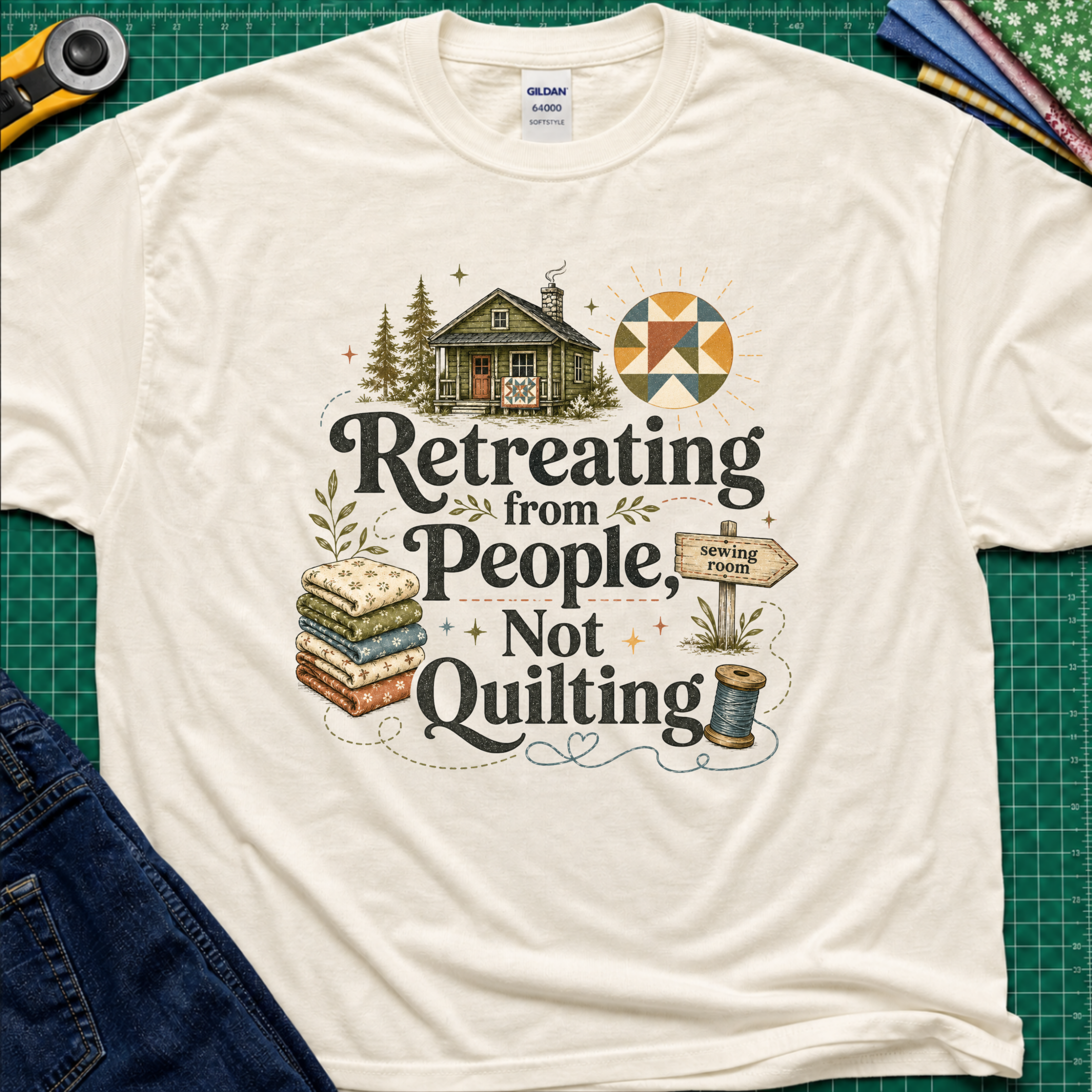 A light cream Gildan t-shirt reading 'Retreating from People Not Quilting' surrounded by illustrations of a cabin, quilts, a sewing machine, and a sign. The shirt is displayed flat on a green cutting mat with quilting tools and jeans partially visible.