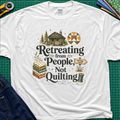 A white Gildan t-shirt reading 'Retreating from People Not Quilting' surrounded by illustrations of a cabin, quilts, a sewing machine, and a sign. The shirt is displayed flat on a green cutting mat with quilting tools and jeans partially visible.