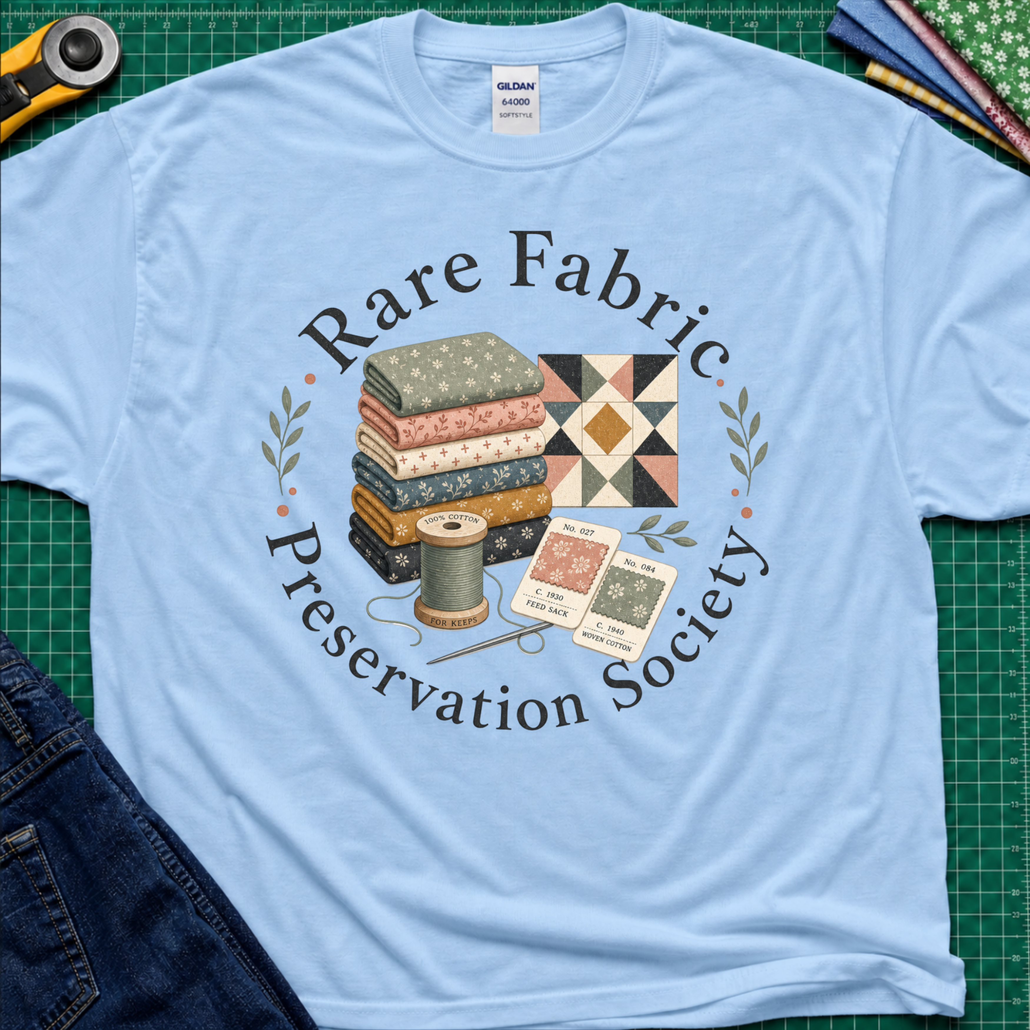 A light blue Gildan t-shirt with a vintage-style graphic reading 'Rare Fabric Preservation Society' around an illustration of stacked quilting fabrics, a spool of thread, and pattern cards. The shirt is laid flat on a green cutting mat with sewing tools and jeans partially visible around it.